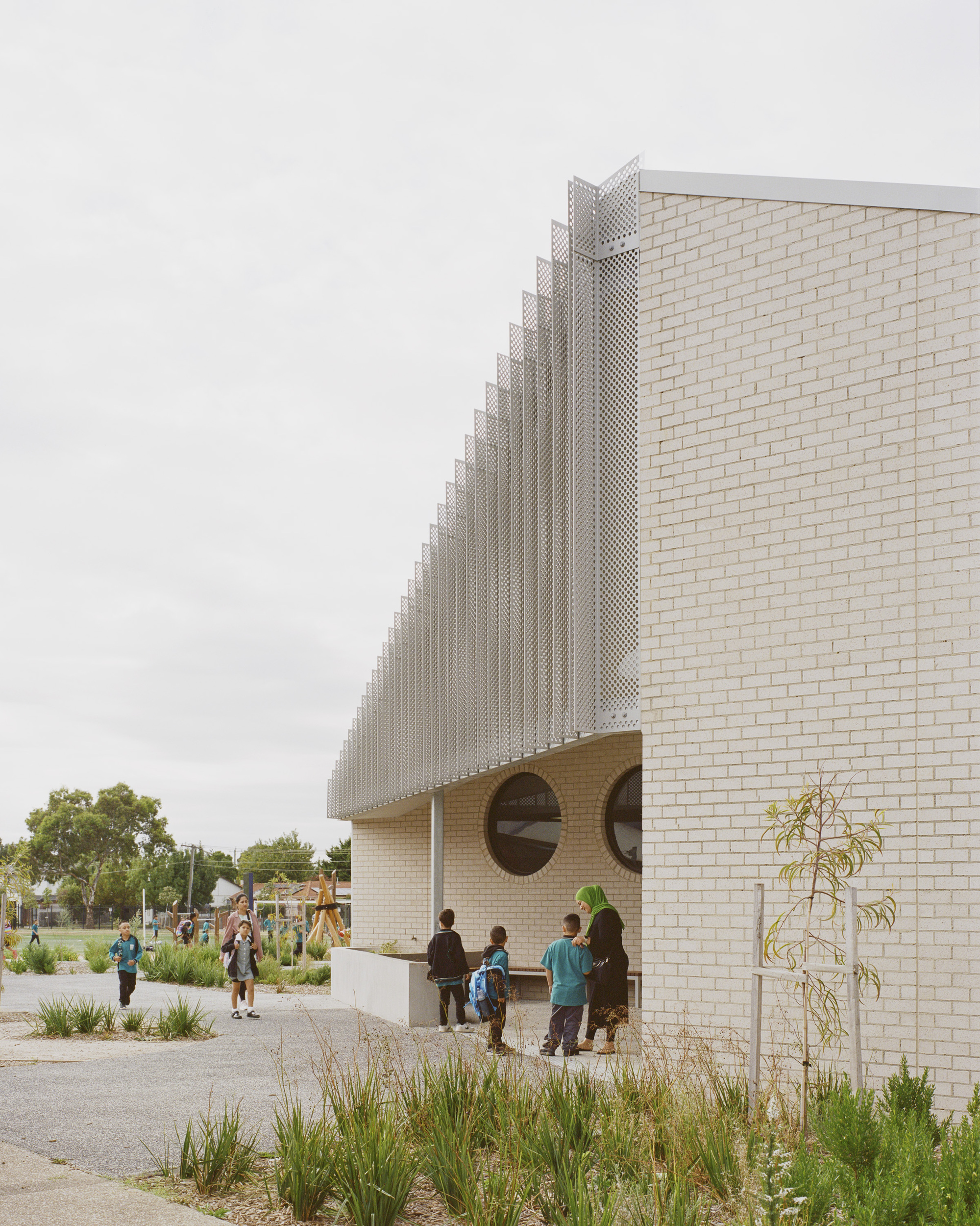 Perforated metal screen rising above pale brick walls with circular openings and visitors gathering beneath an overcast sky