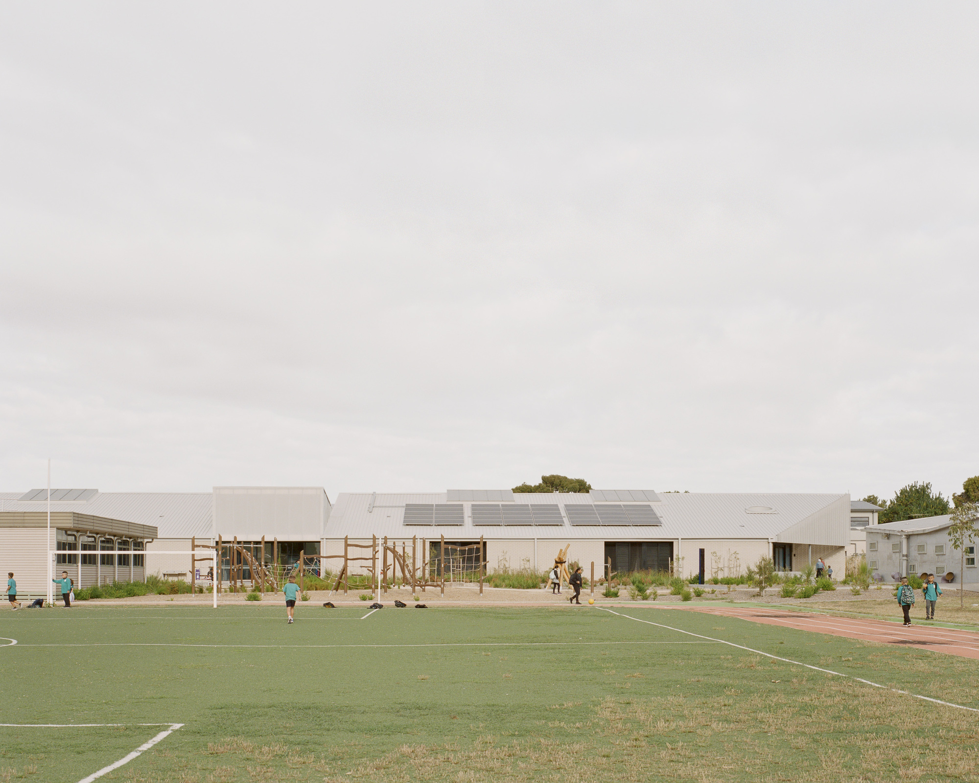 View across sports field showing low-slung building with solar panels on sawtooth roofs and children playing on grass