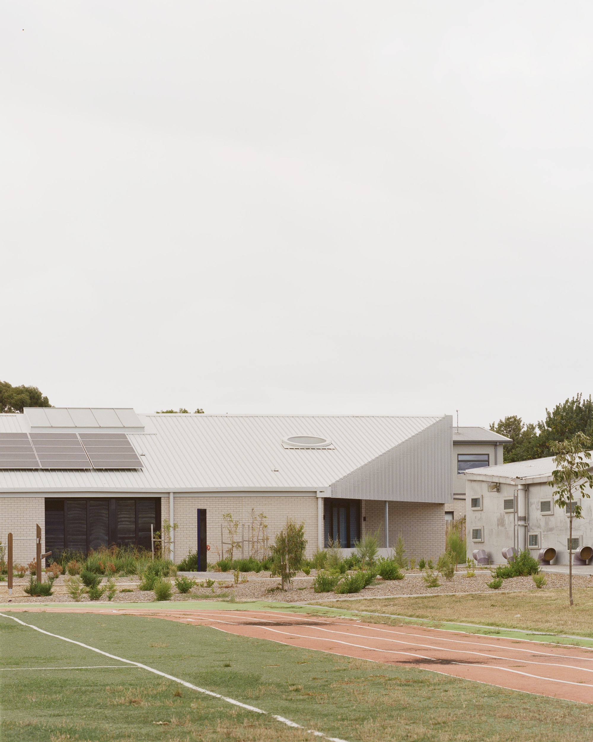 Corrugated metal roof volumes with solar panels fronting a planted native garden and running track under grey skies