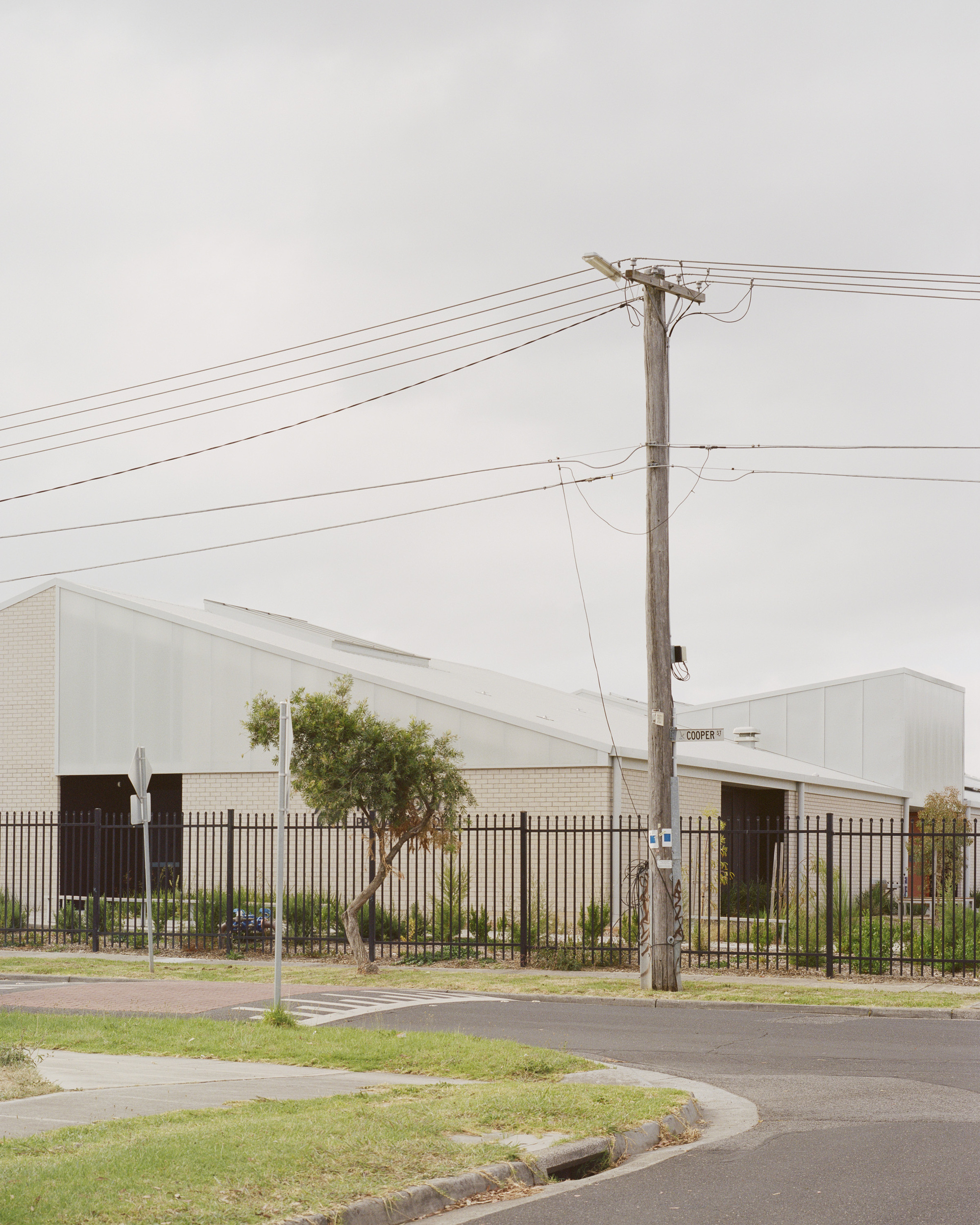 Street view showing pale brick and metal roofed volumes behind security fence with utility pole in foreground