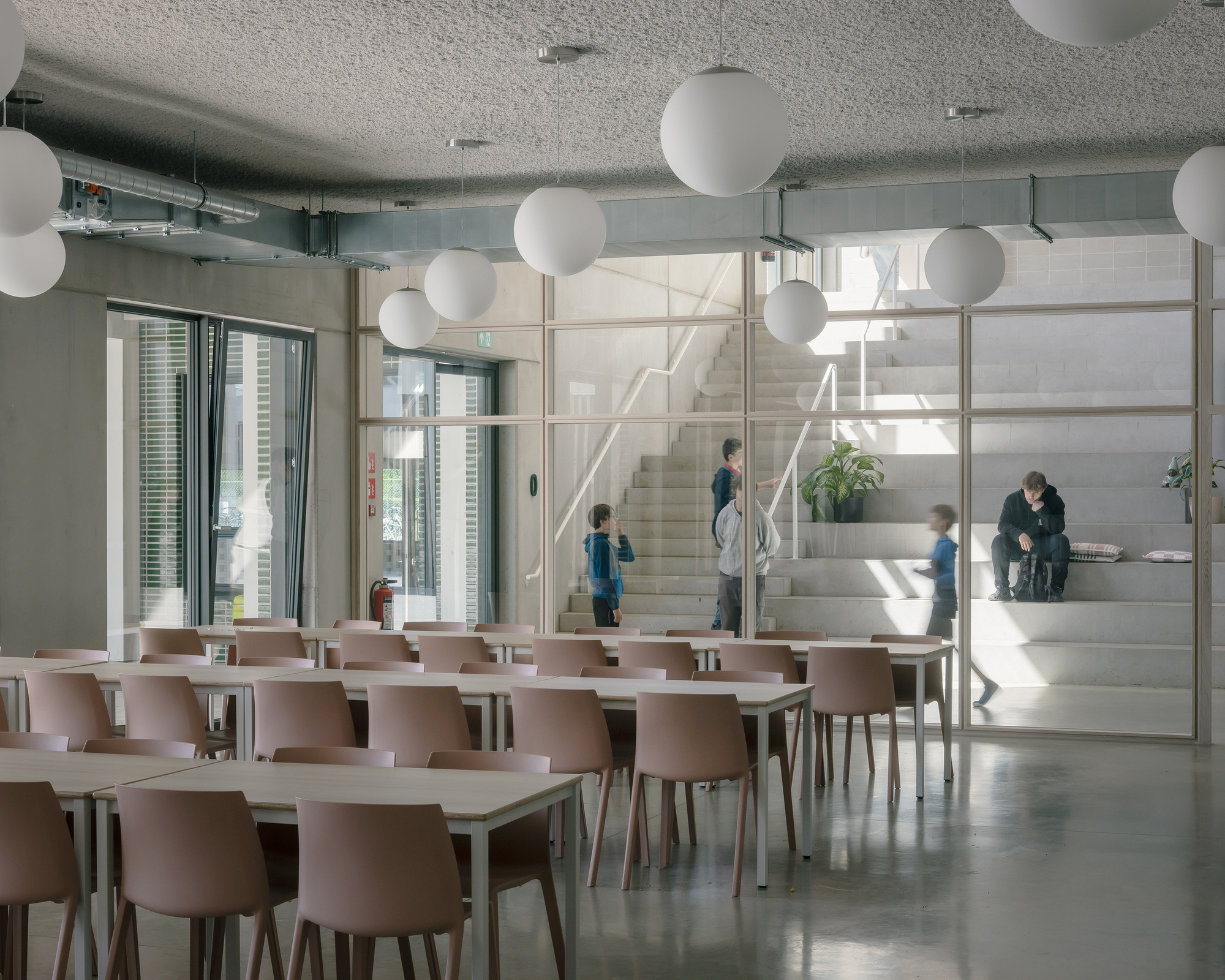 Dining hall with pale pink chairs and globe pendant lights overlooking a glass-enclosed stairwell