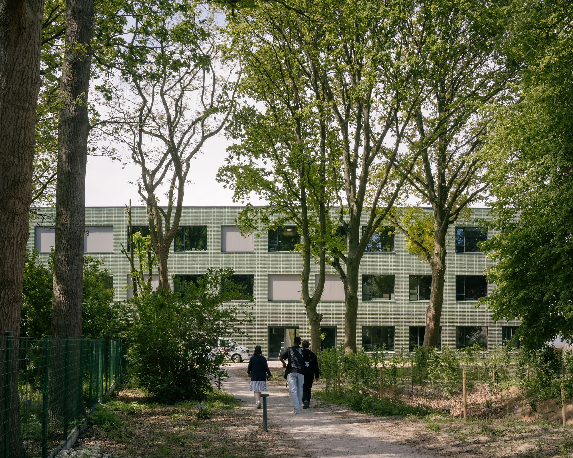 Green tiled facade viewed through a tree-lined path with pedestrians walking towards the entrance