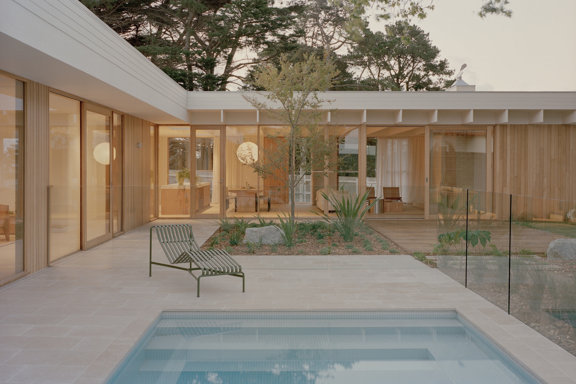 Courtyard pool with glass fencing and planted bed surrounded by timber framed glazing at dusk