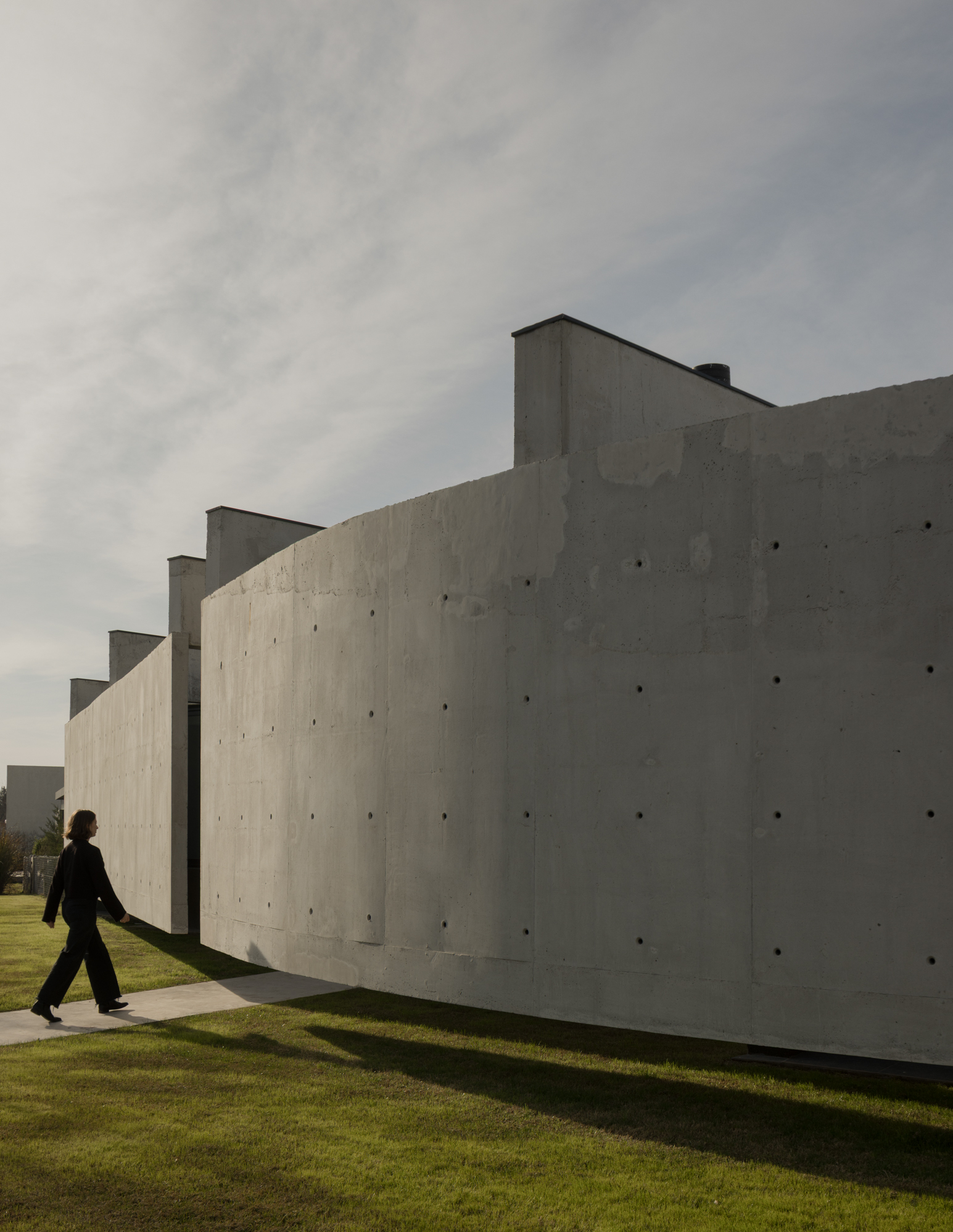 Stepped concrete facade with visible formwork ties as a figure walks along the pathway