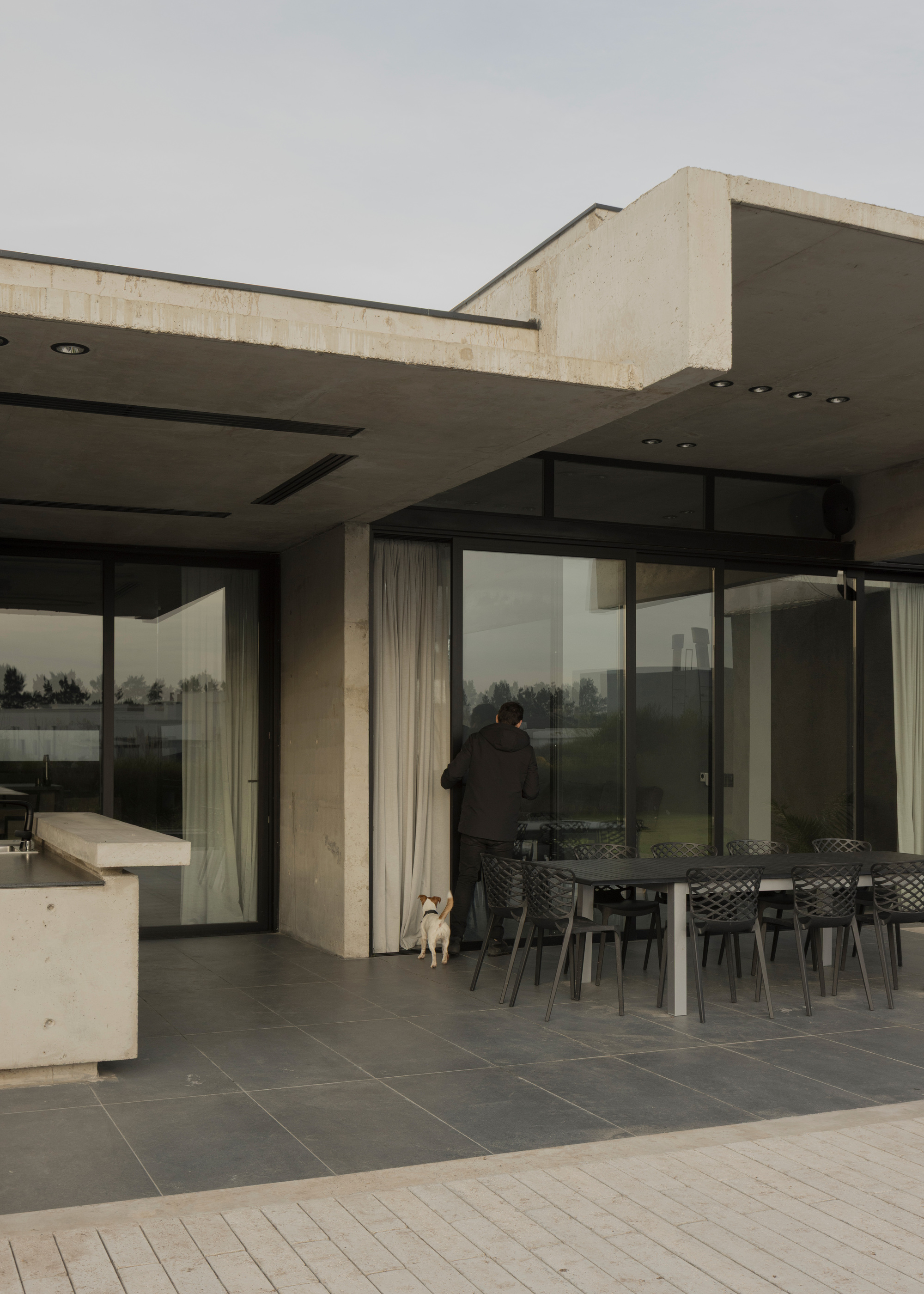 Covered outdoor terrace with dining table, concrete beams, and a dog at the doorway