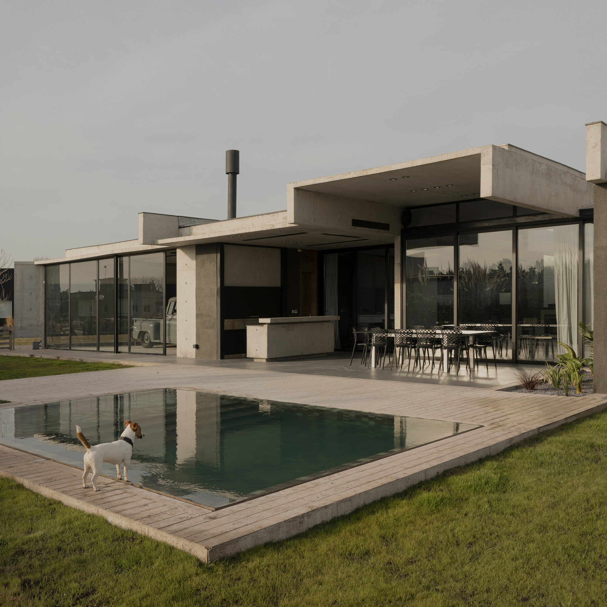 Pool deck with timber edging, glazed pavilion, outdoor kitchen, and a dog by the water