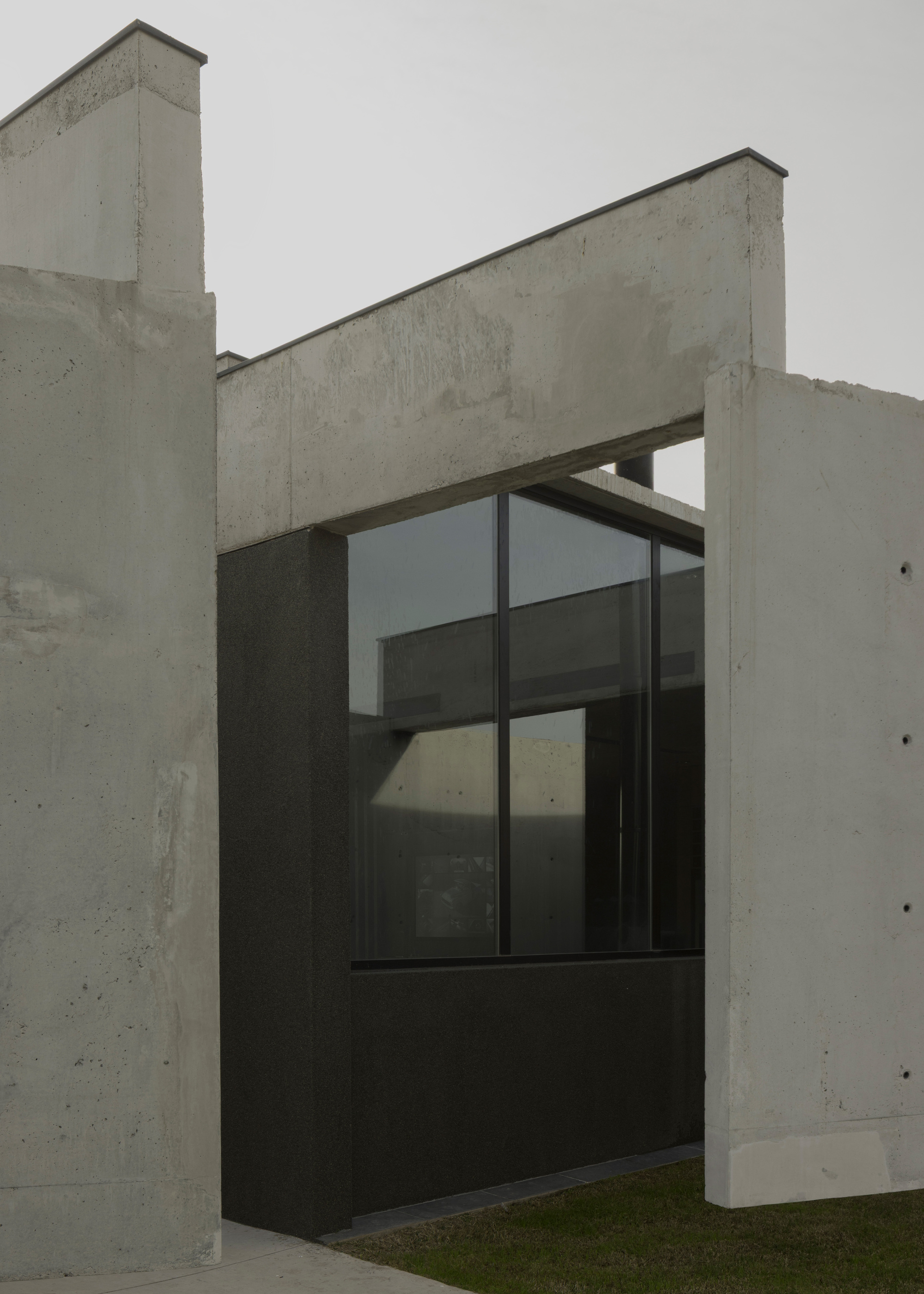 Corner detail of concrete volumes framing a projecting black-framed window bay under overcast sky