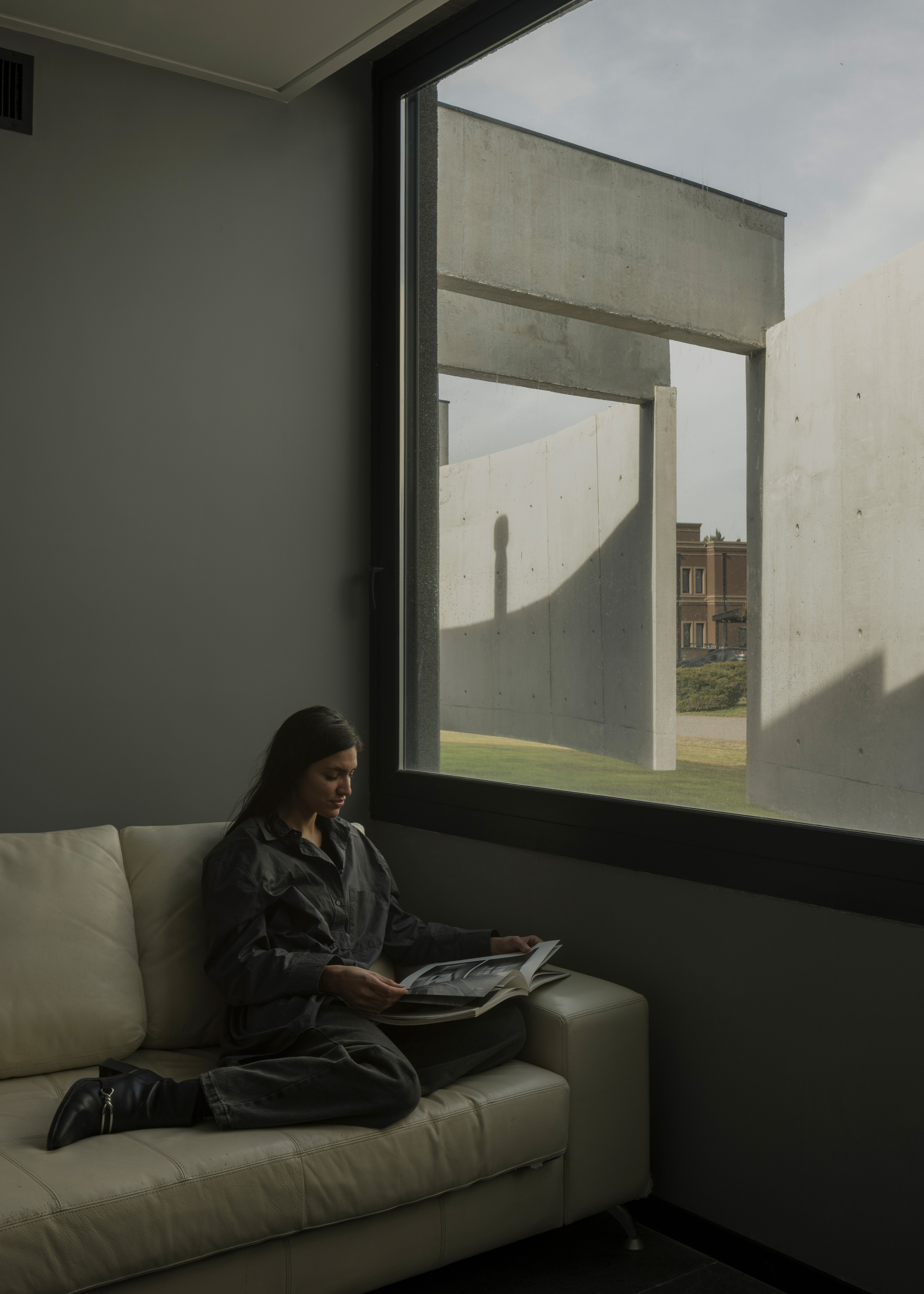 Interior reading nook with woman on sofa beside a deep-set window framing concrete courtyard walls
