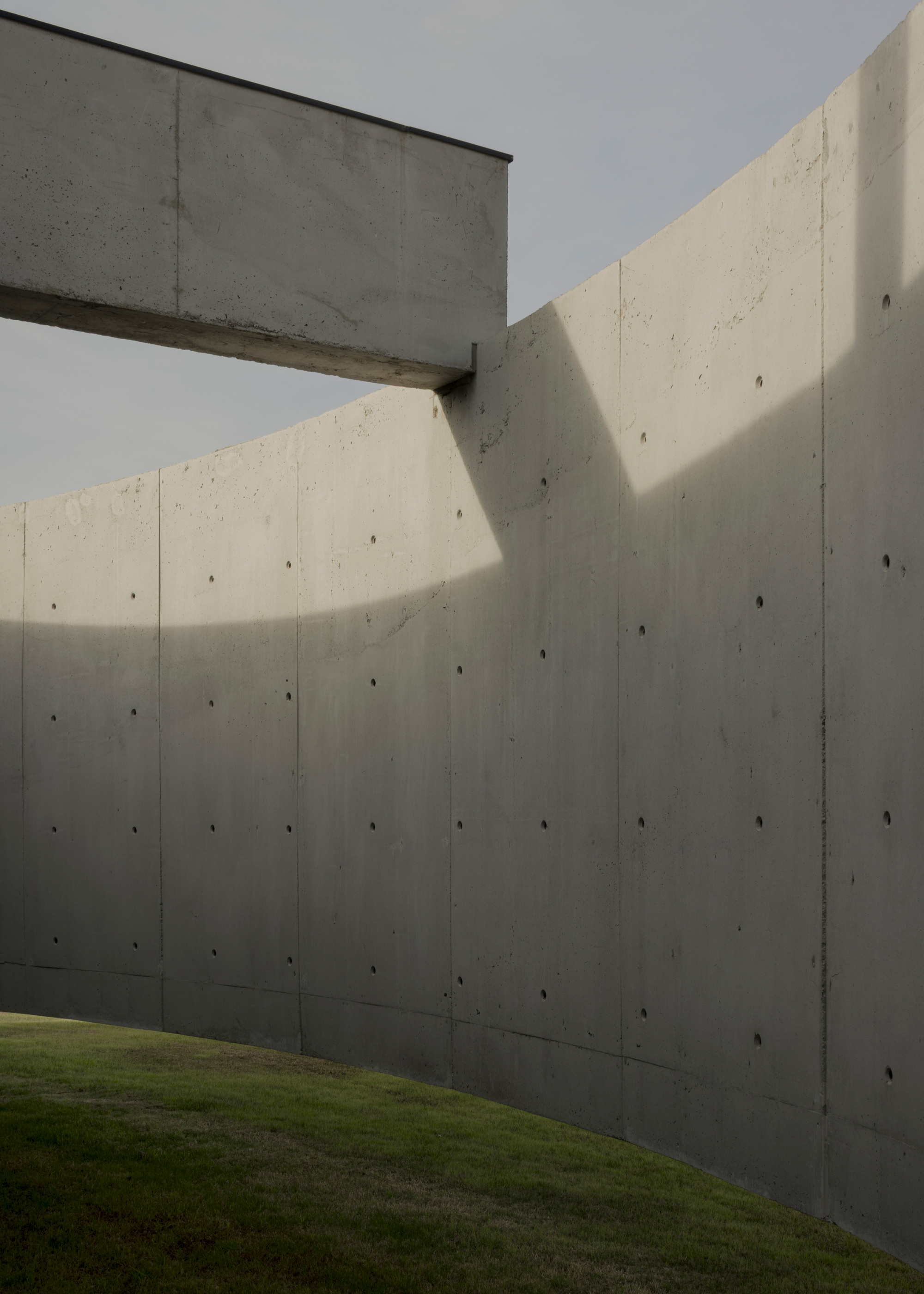 Boardformed concrete walls meeting at an angled corner with a cantilevered beam above green lawn