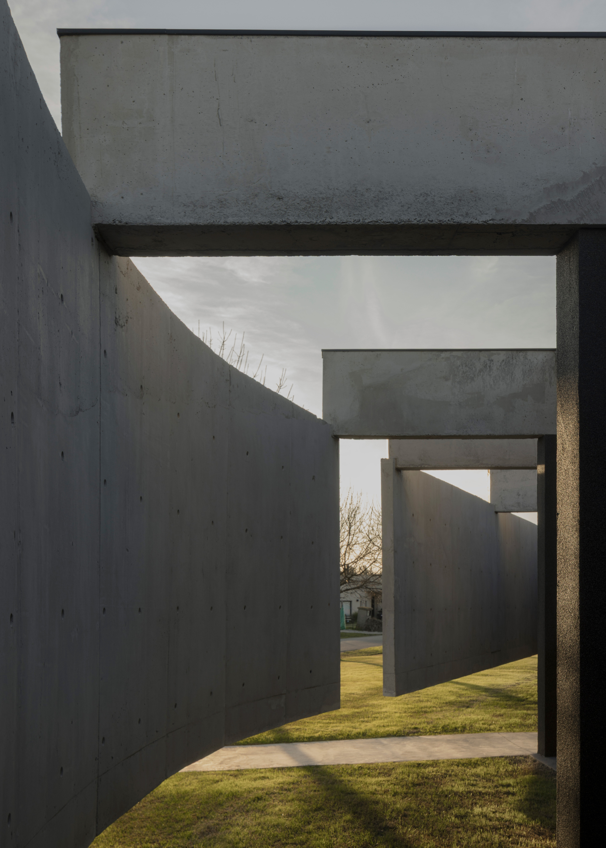 Passageway between concrete walls creating layered frames looking toward lawn and bare trees at dusk