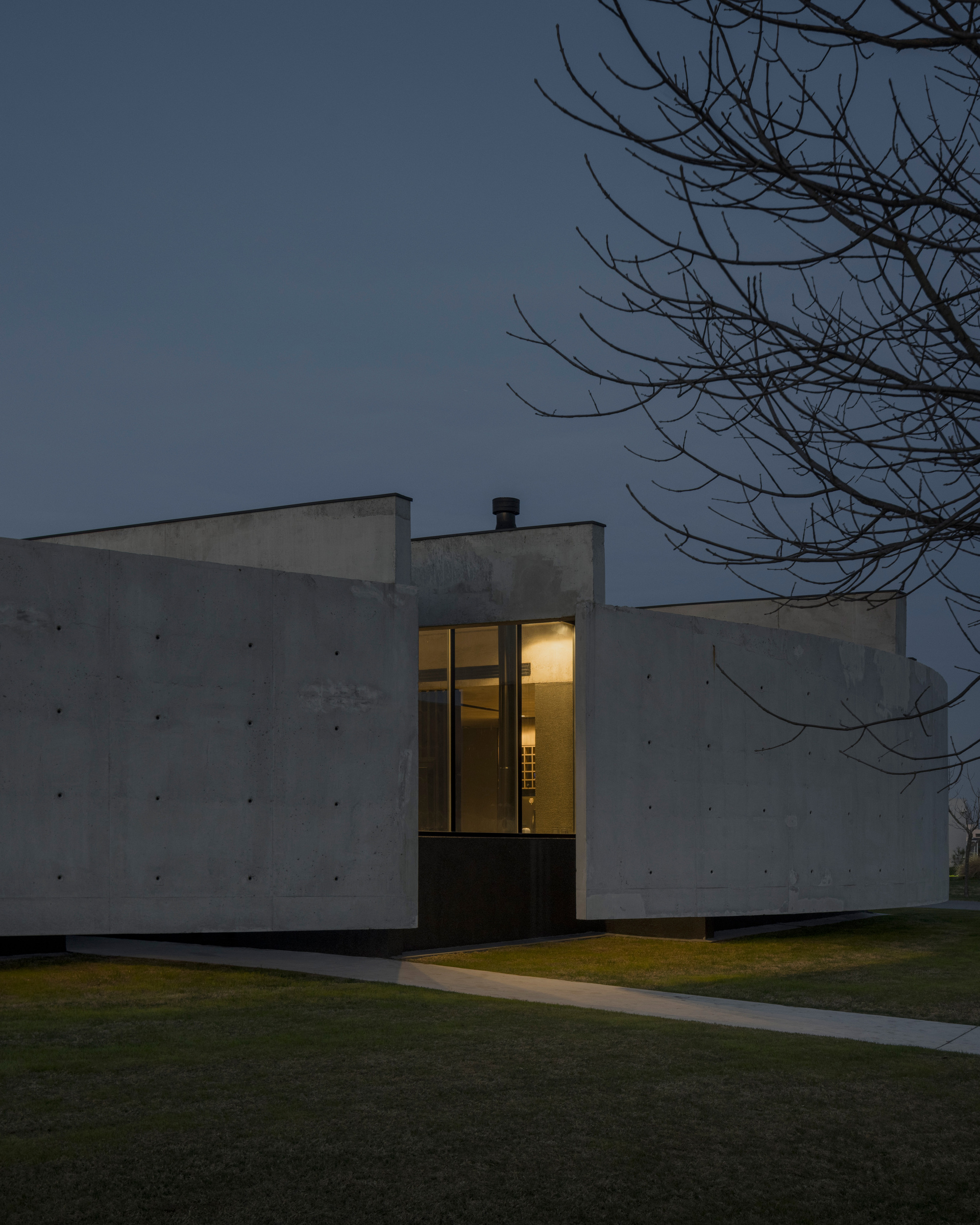 Dusk view of the illuminated corner window within the board-formed concrete facade beneath bare branches