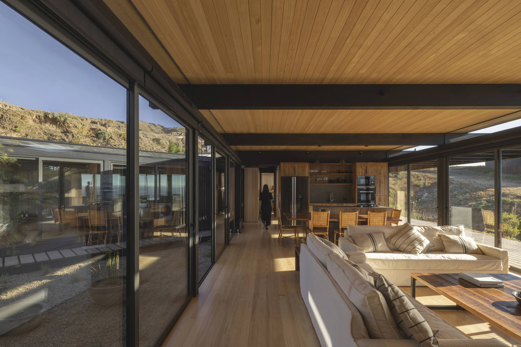 Open-plan living space with timber ceiling, exposed steel beams and floor-to-ceiling glass overlooking dry hillside