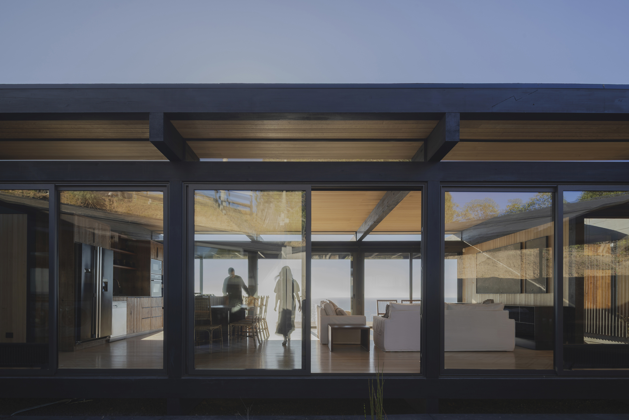 Exterior view through glazed facade showing interior spaces with timber ceiling and figures moving at dusk