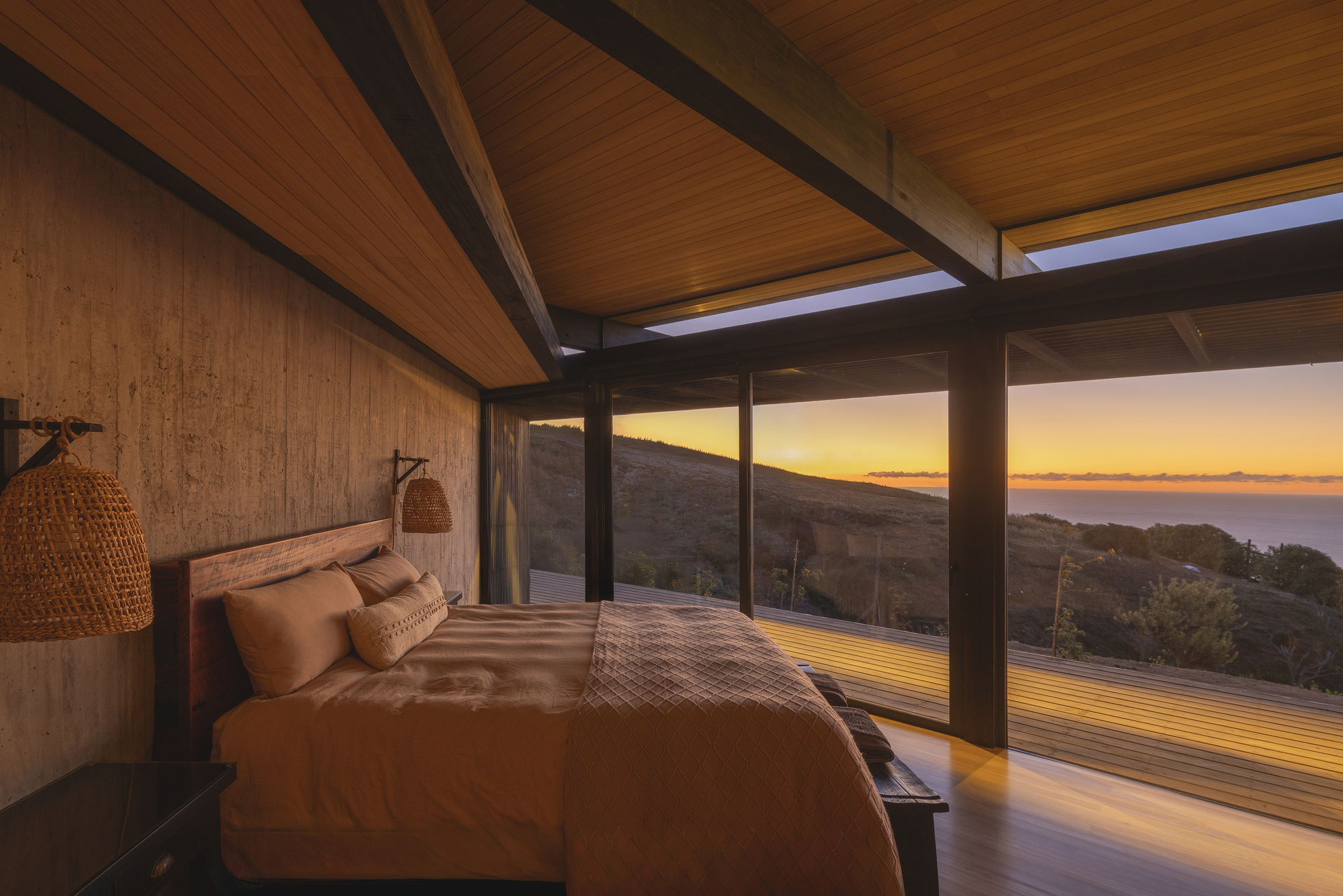 Bedroom with timber walls and ceiling framing floor-to-ceiling windows overlooking the coastline at sunset