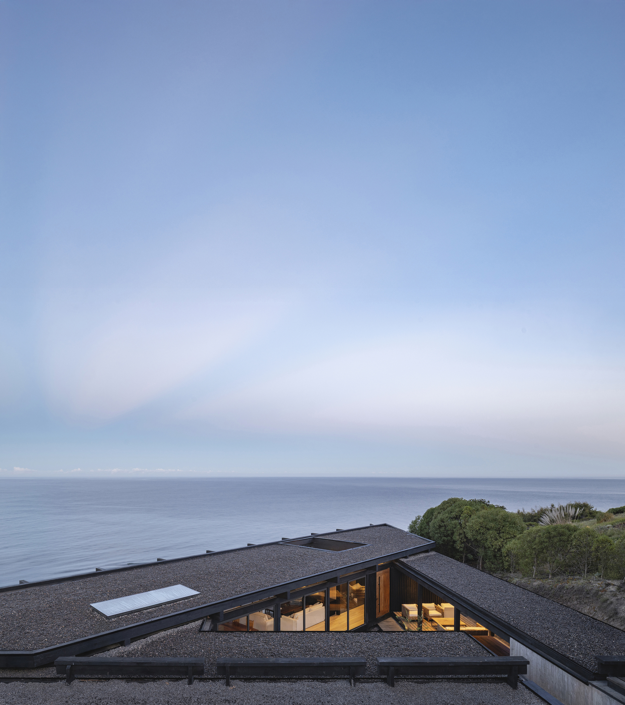 Angled gravel roof with skylights and glazed openings set into coastal vegetation at dusk