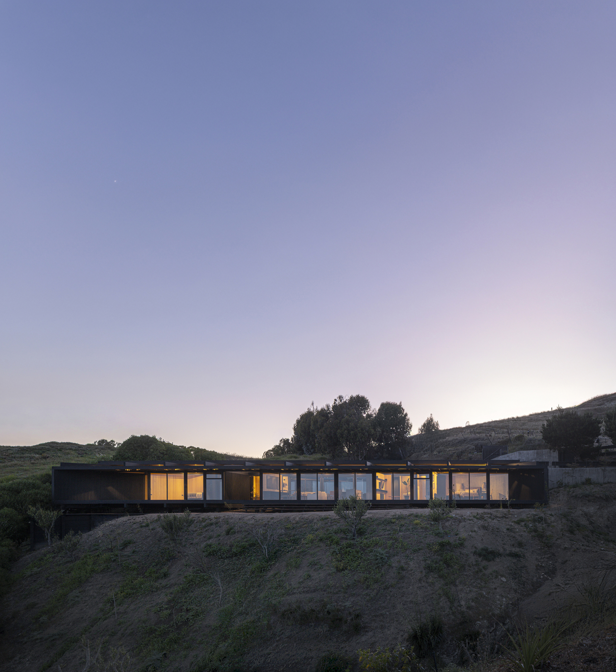 Illuminated linear volume set into a hillside viewed at dusk with rolling terrain and trees beyond