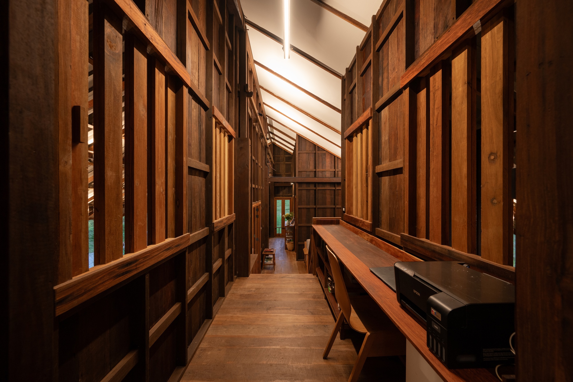 Interior corridor with timber-framed shelving walls and skylight casting striped shadows across the wooden floor