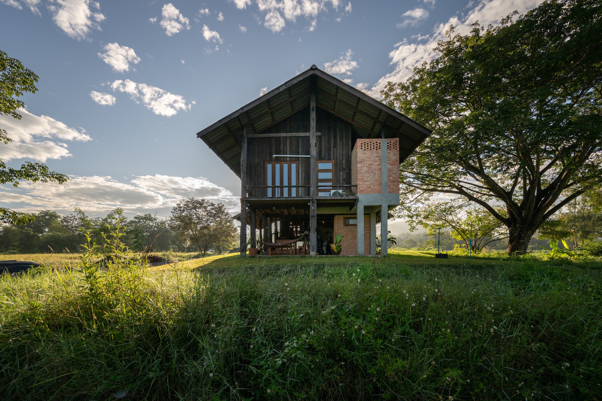 Two-story house raised on timber stilts over a grass field under afternoon clouds