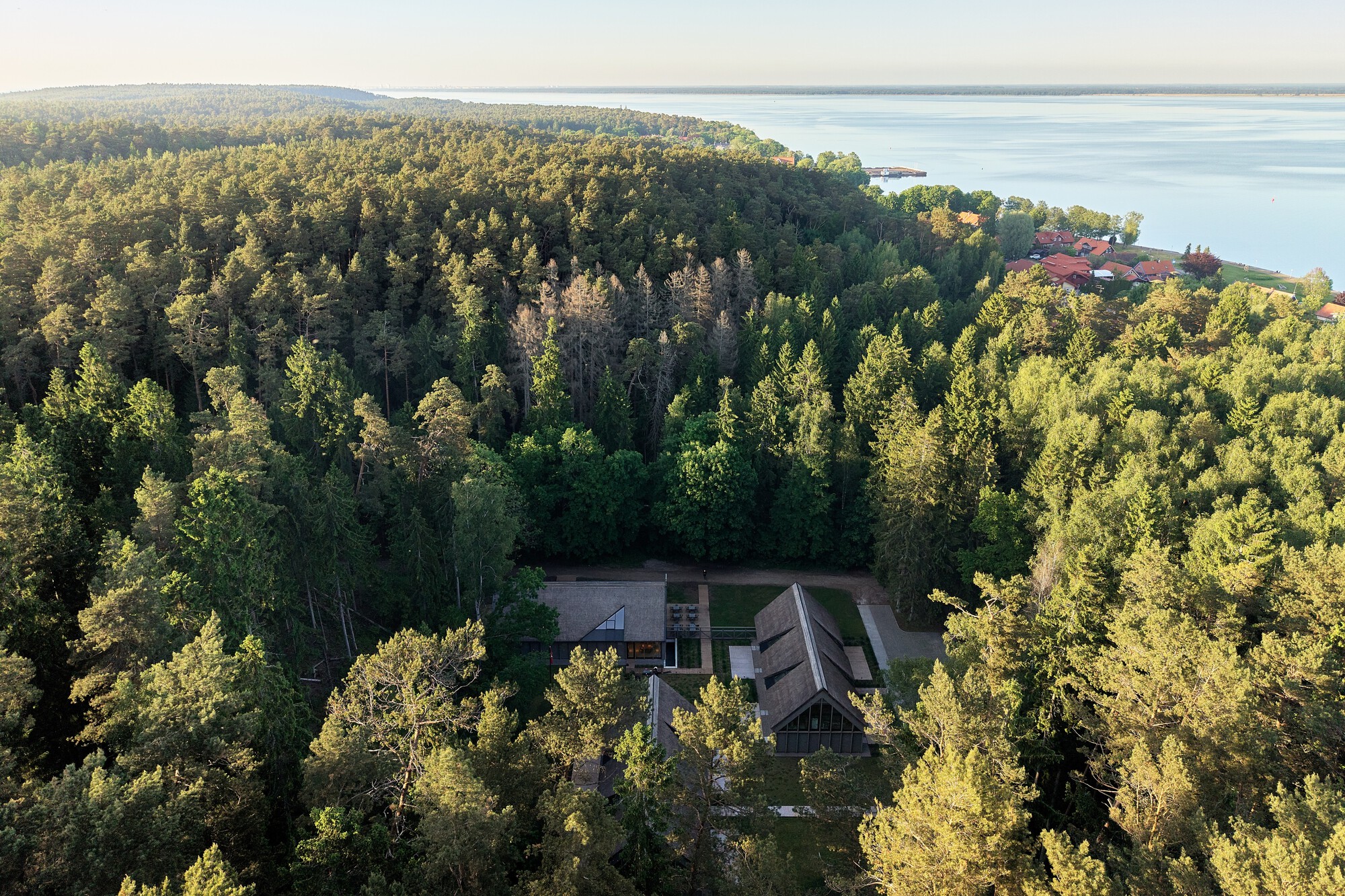 Aerial view of three gabled volumes nestled in a forest clearing near a lakeshore