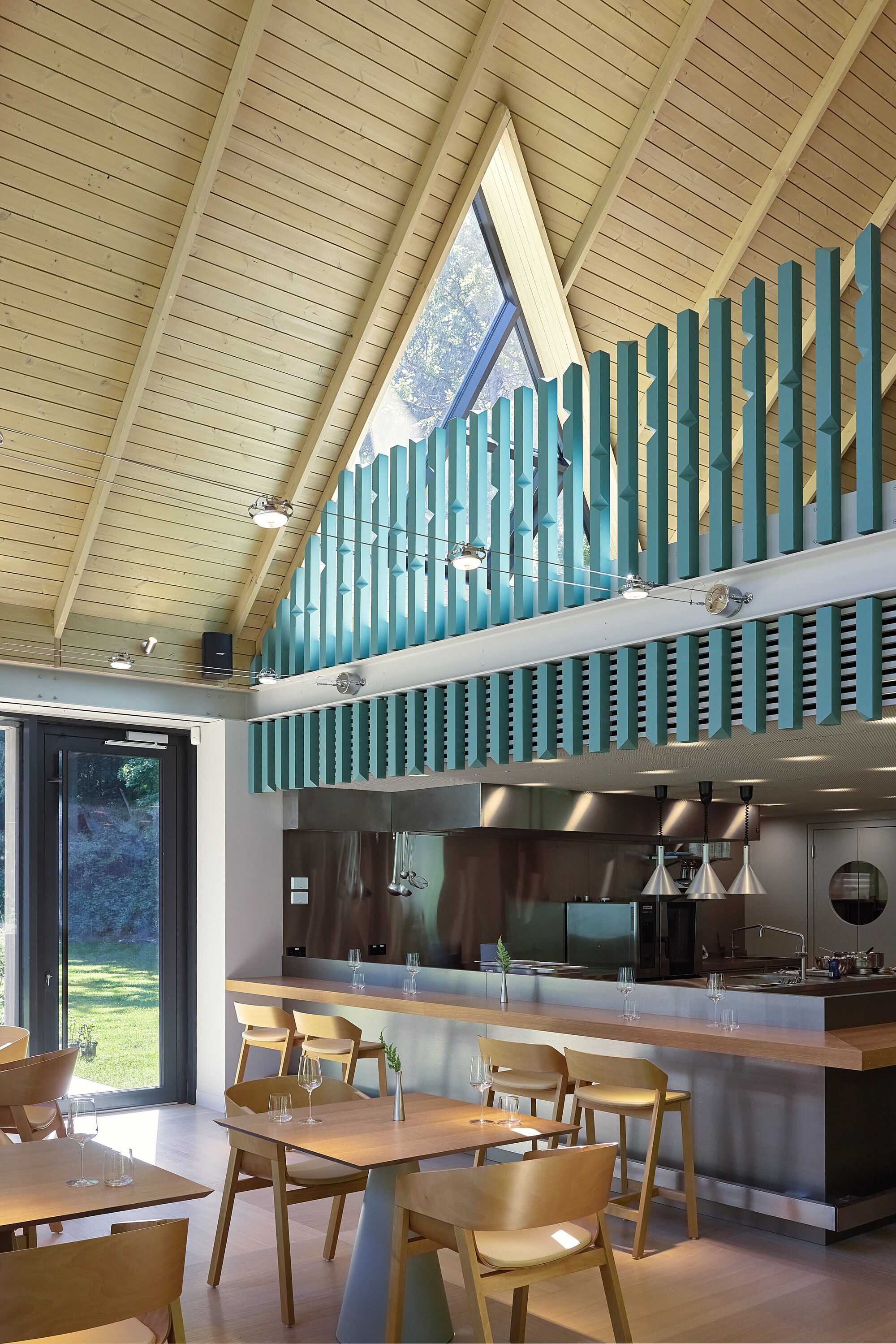 Dining hall interior with timber-lined vaulted ceiling, vertical teal slat screen above the steel kitchen counter