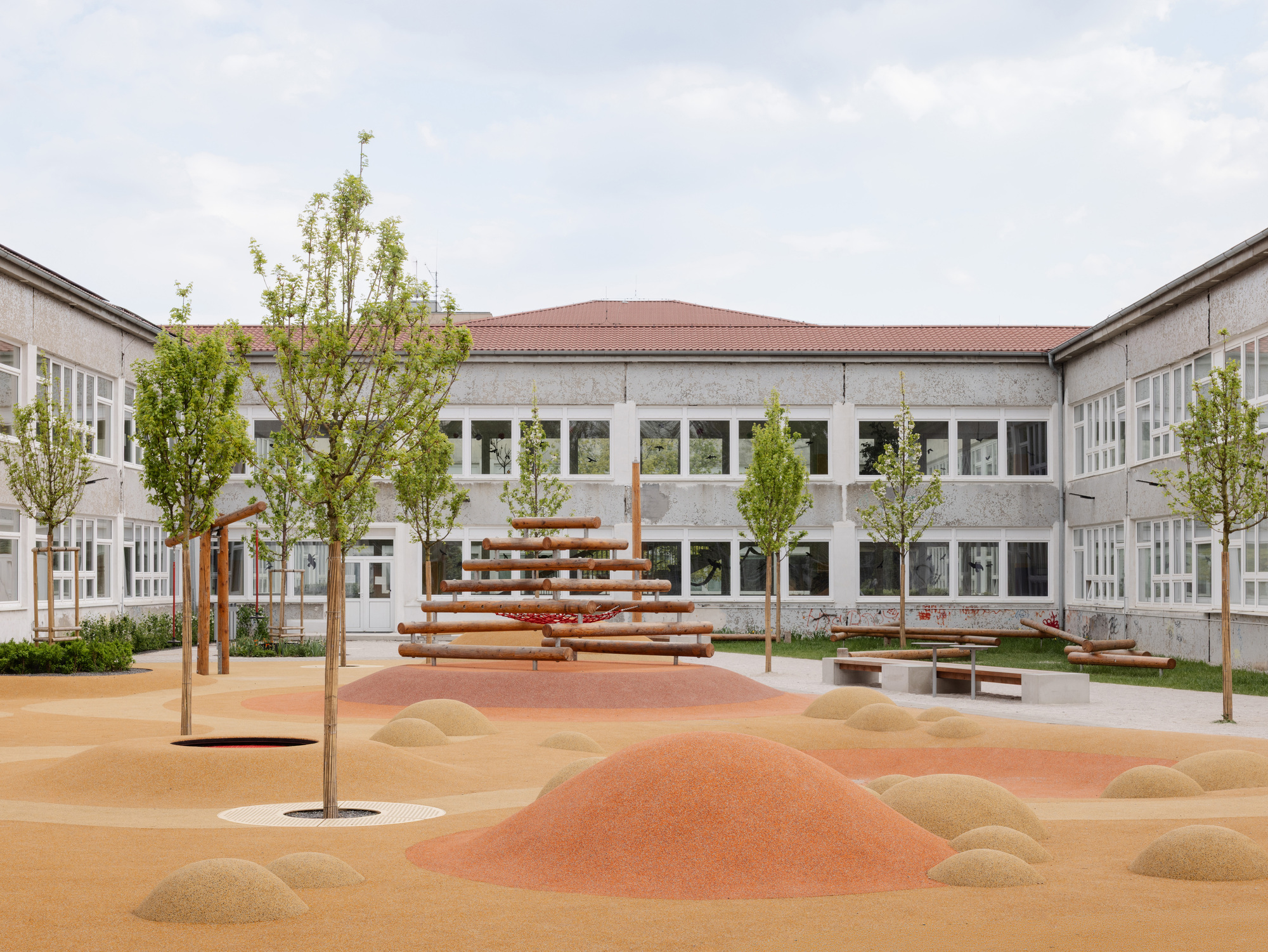 Courtyard playground with sculpted mounds and young trees beneath cloudy skies
