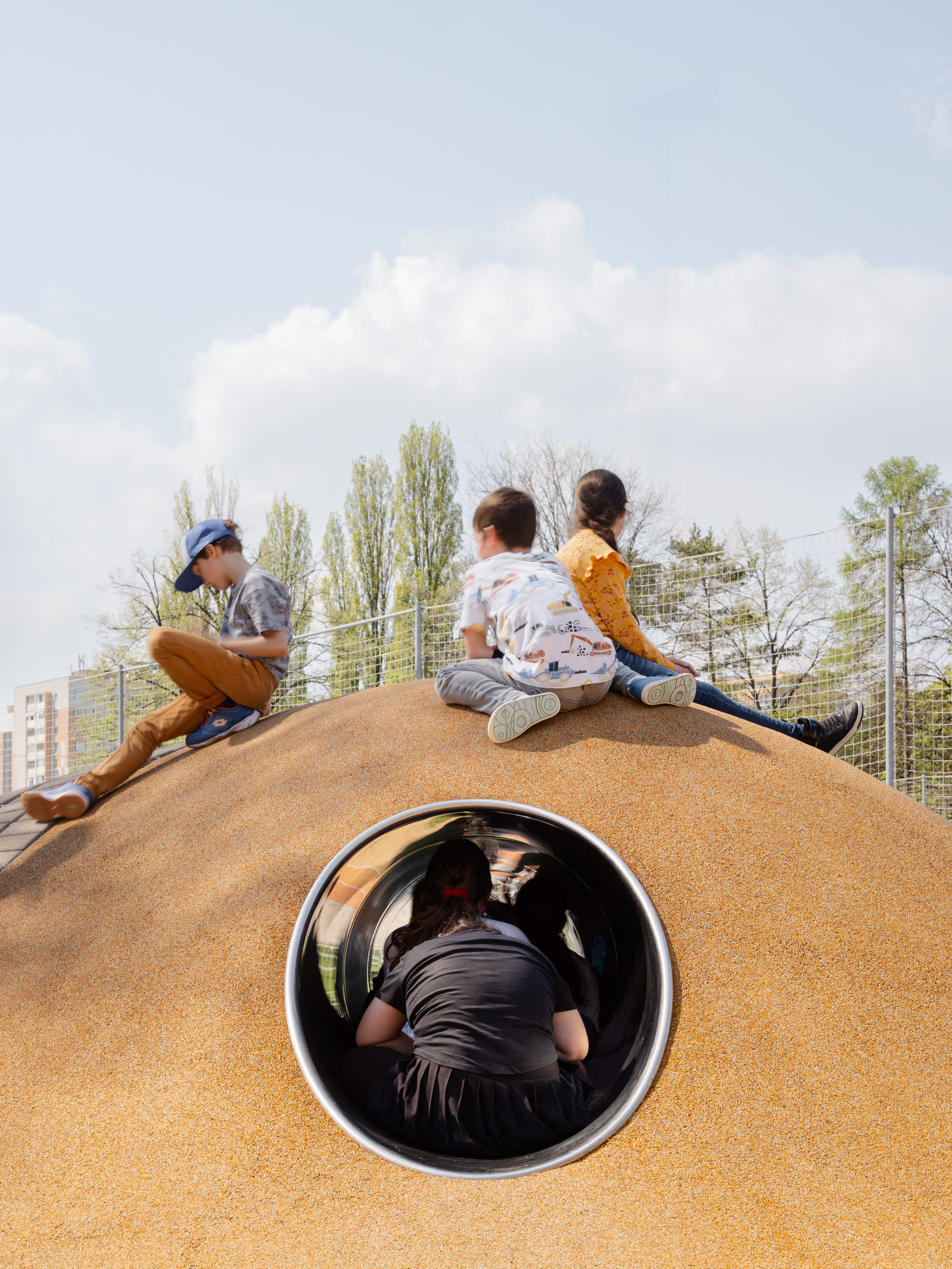 Children sitting on orange mound above black tubular tunnel opening under cloudy sky