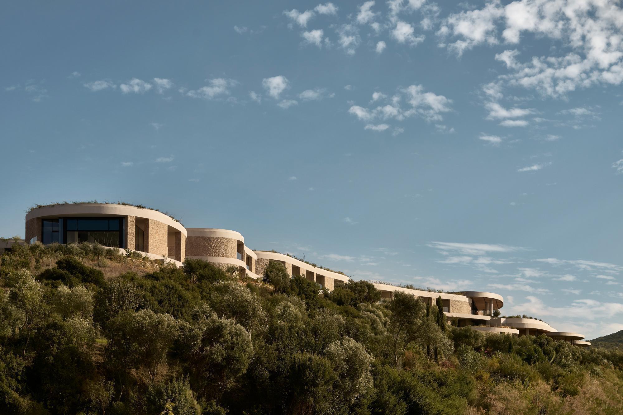 Curving stone-clad volumes nestled into a hillside covered with scrub vegetation under scattered clouds
