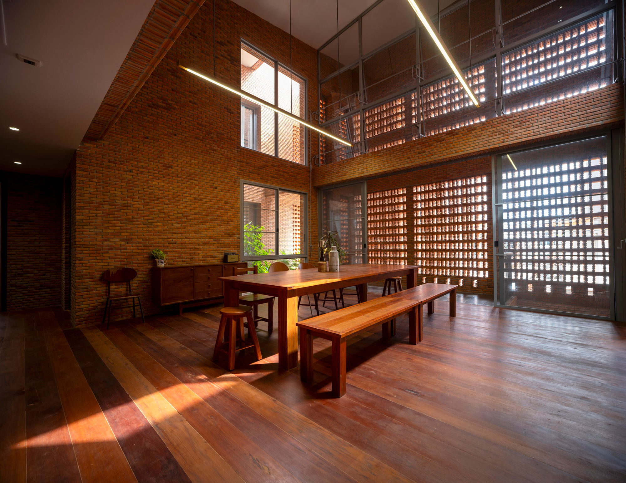Double-height dining space with timber table, benches, and perforated brick screens filtering daylight