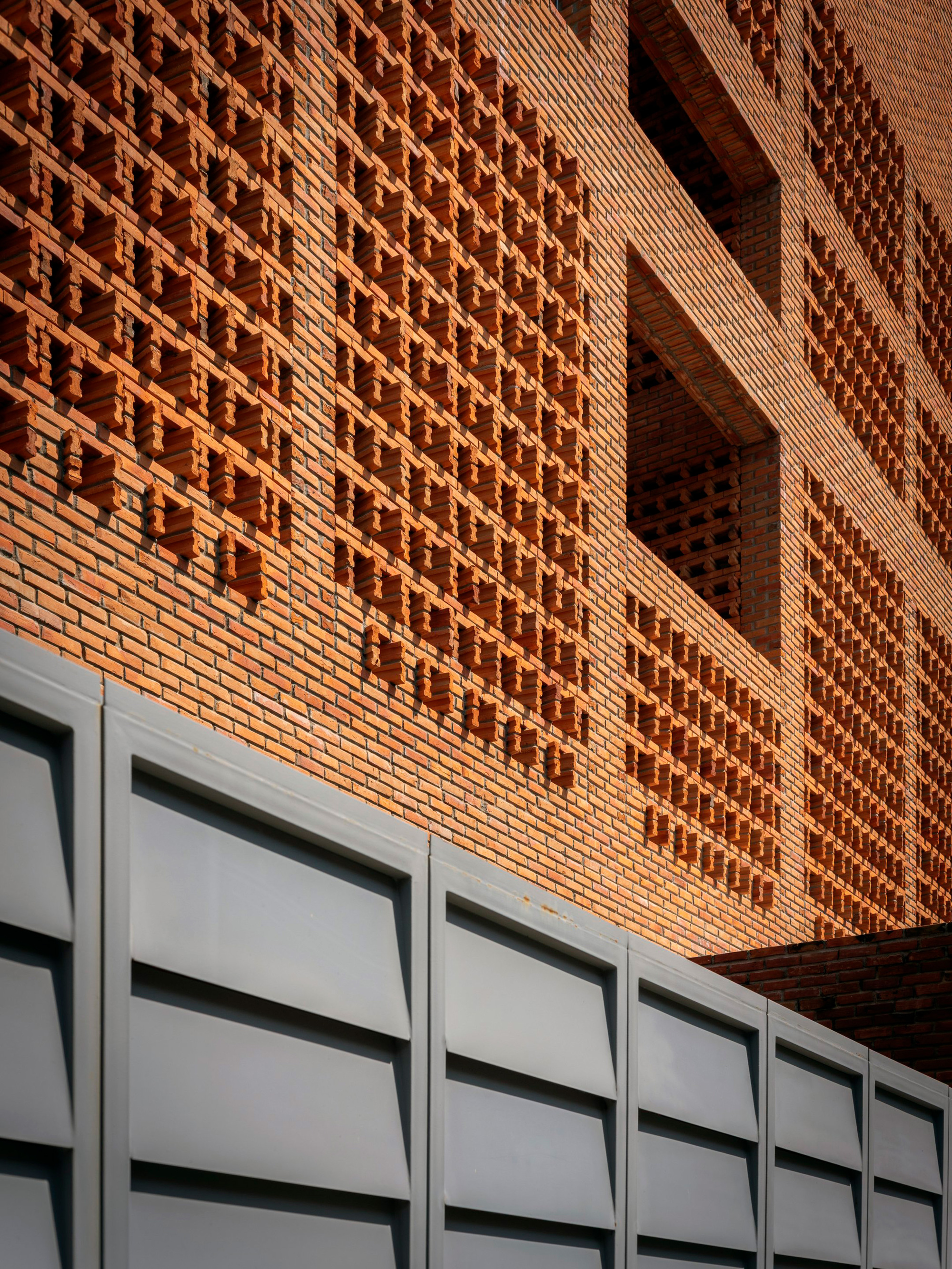 Textured brick facade with alternating solid and perforated patterns above white metal paneling in afternoon light