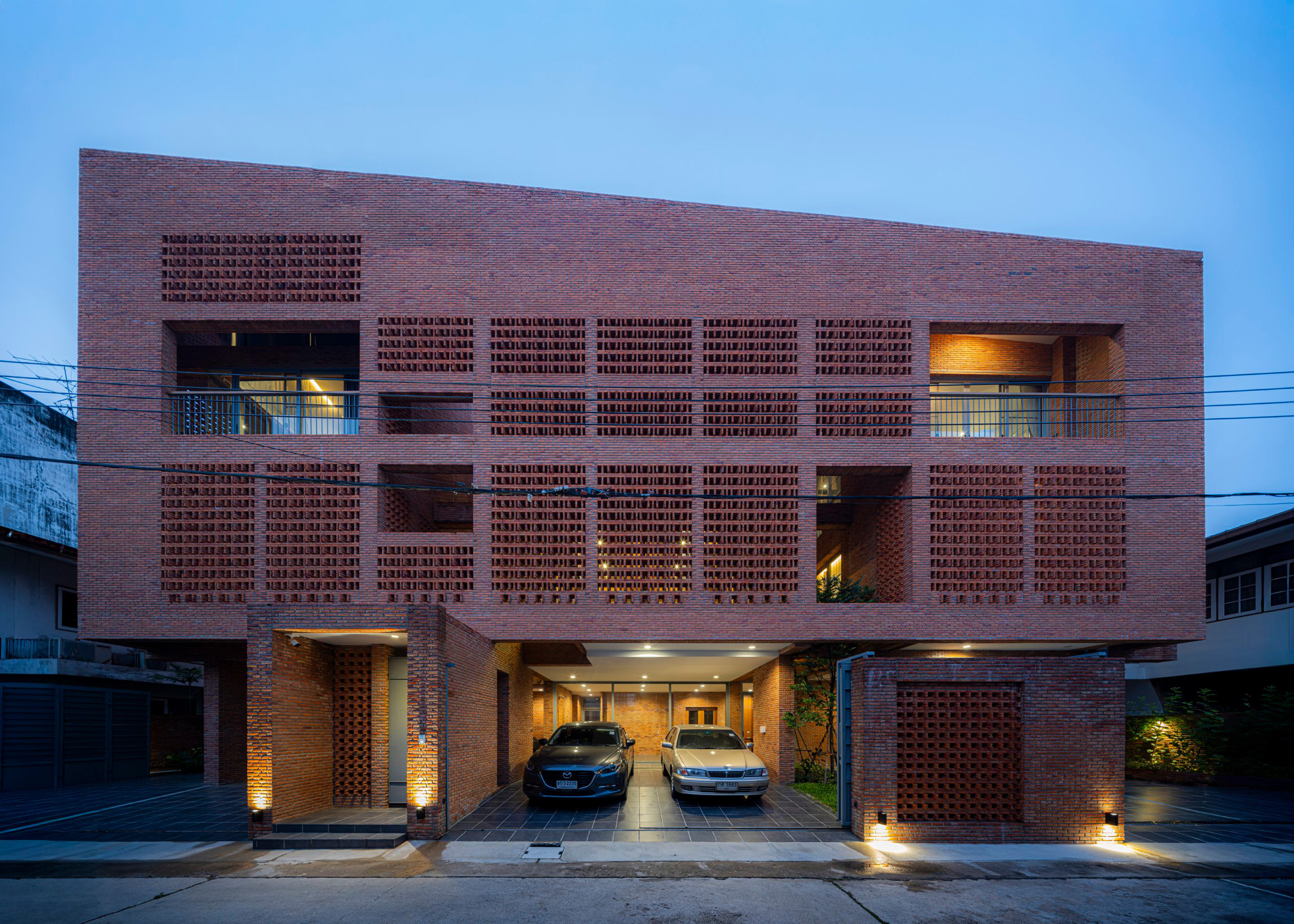 Street facade with perforated brick screens and illuminated openings at dusk revealing parked cars inside
