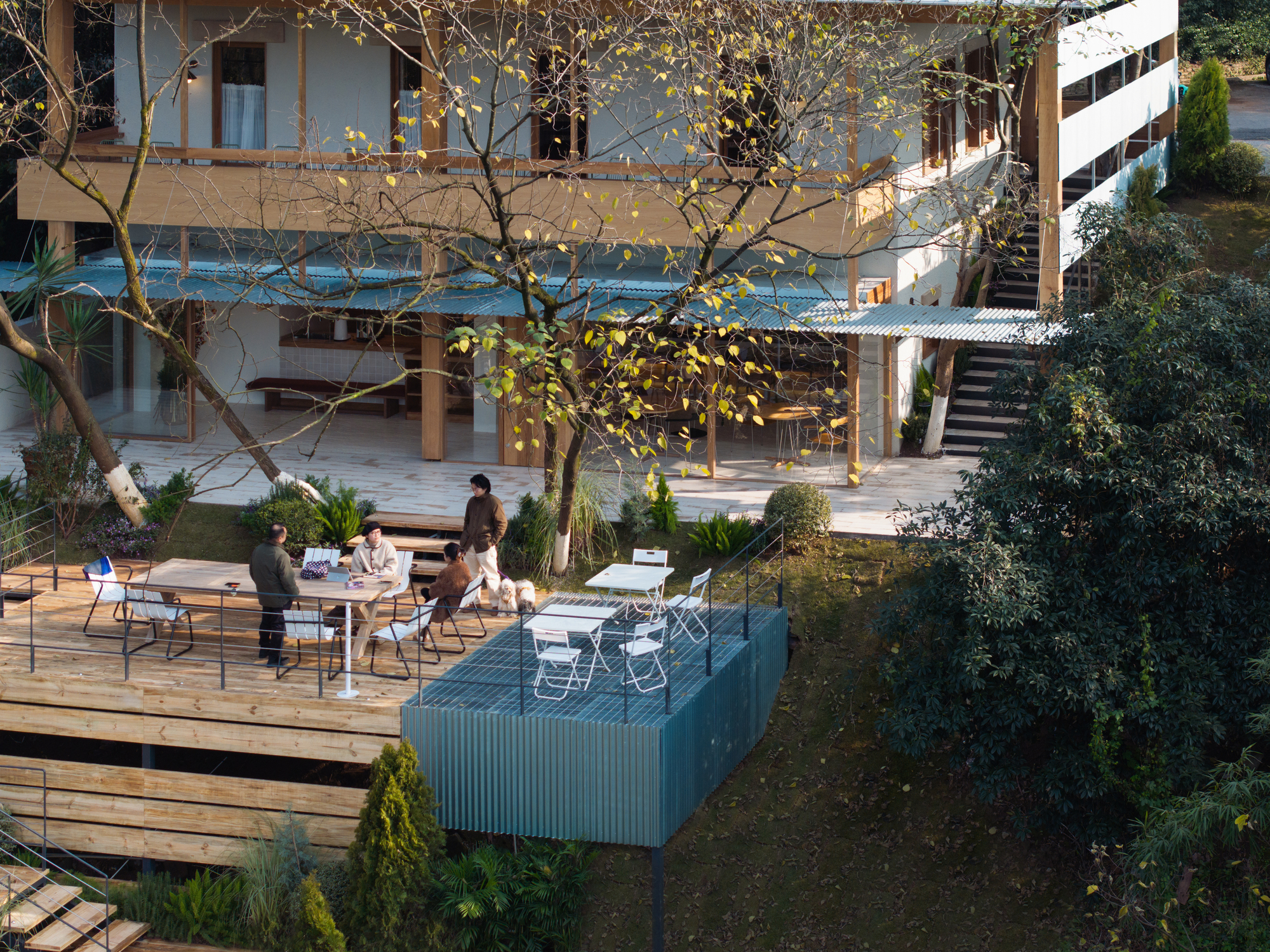 Outdoor terrace with timber decking and corrugated metal volume where figures gather around tables in afternoon light