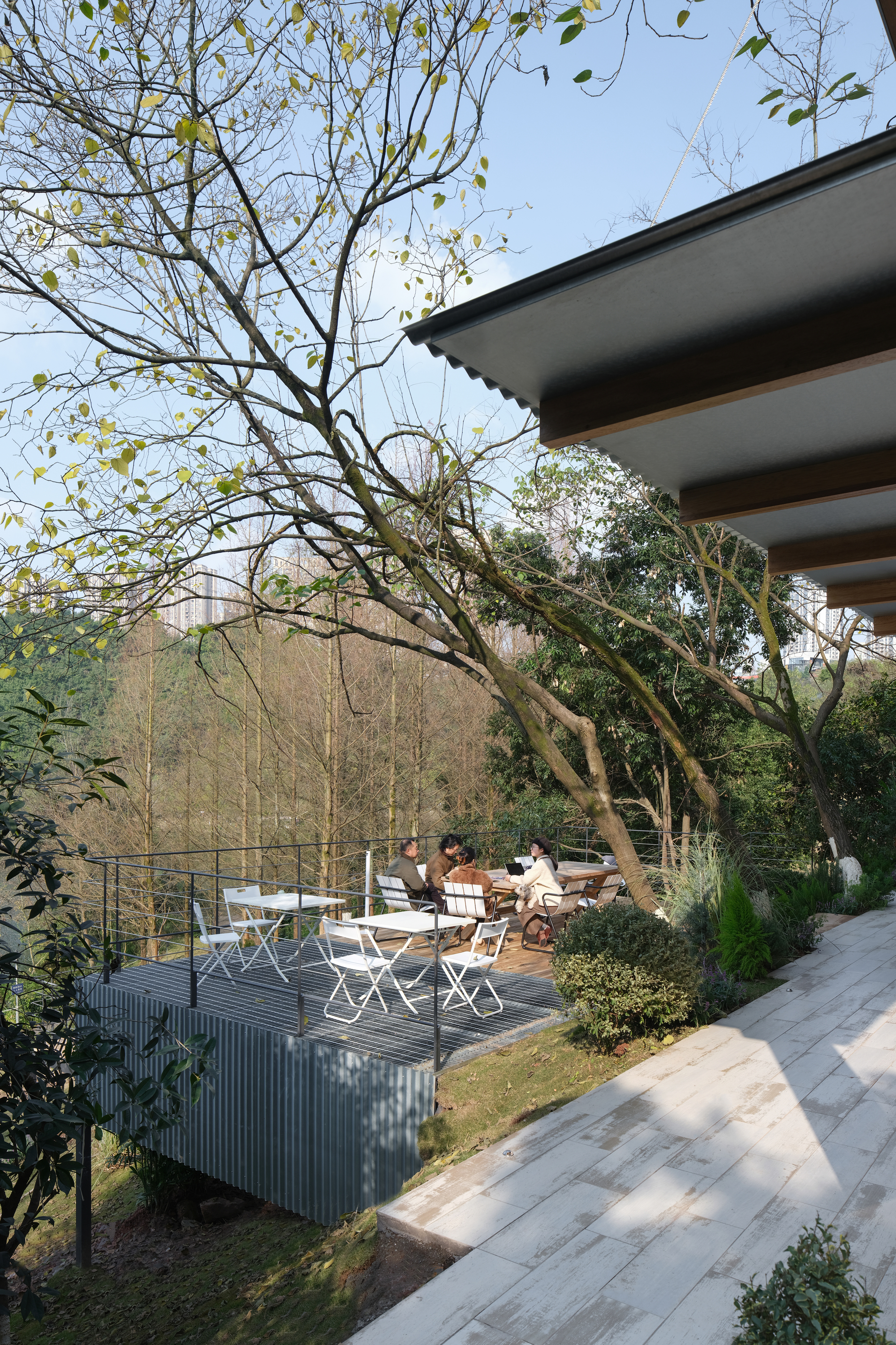 Garden terrace with corrugated metal planter and white folding chairs beneath deciduous trees