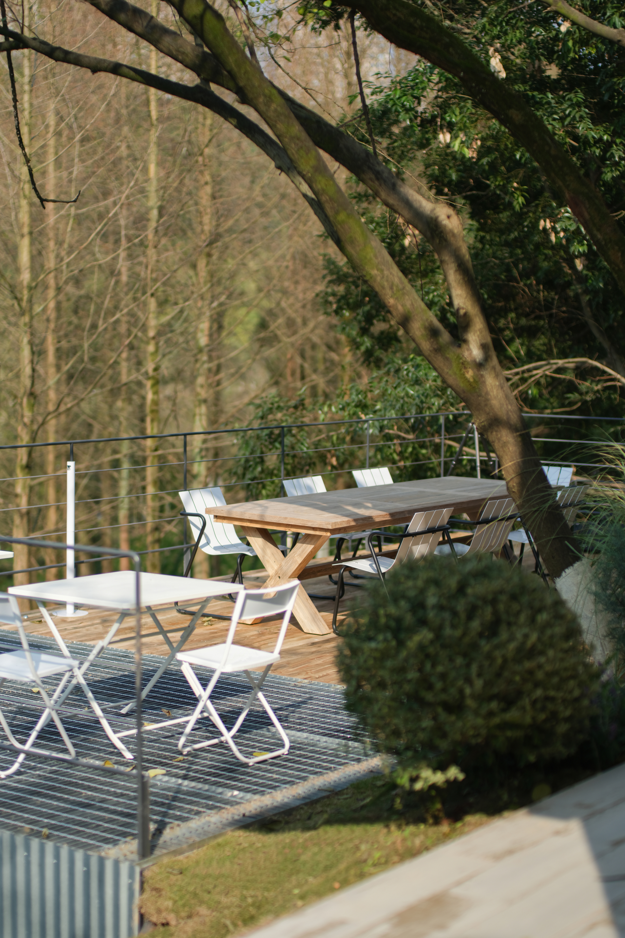 Outdoor dining area with timber table and folding chairs on metal grating deck beneath tree canopy