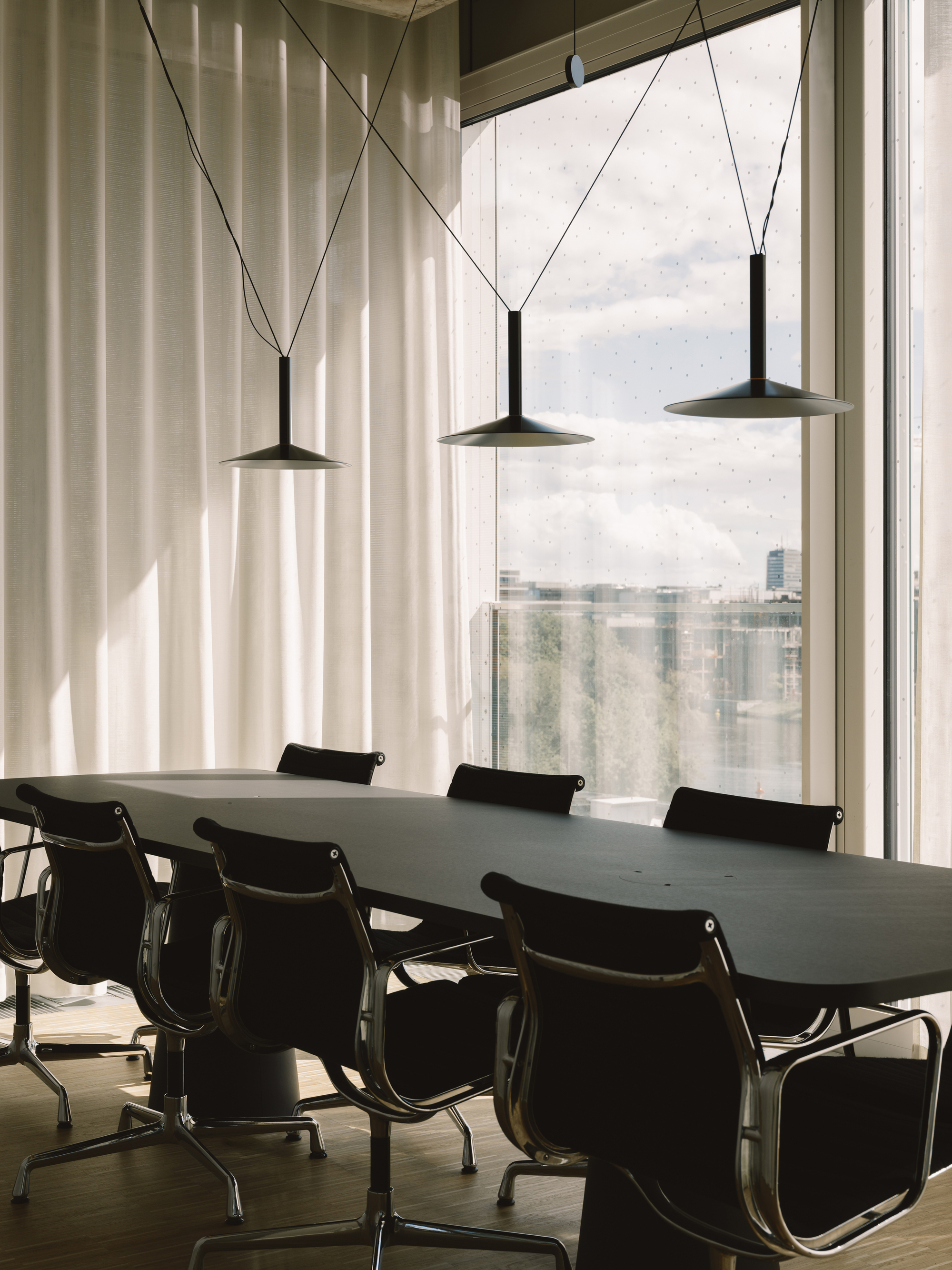 Conference table with leather chairs beneath three pendant lights suspended by cables near a city-view window