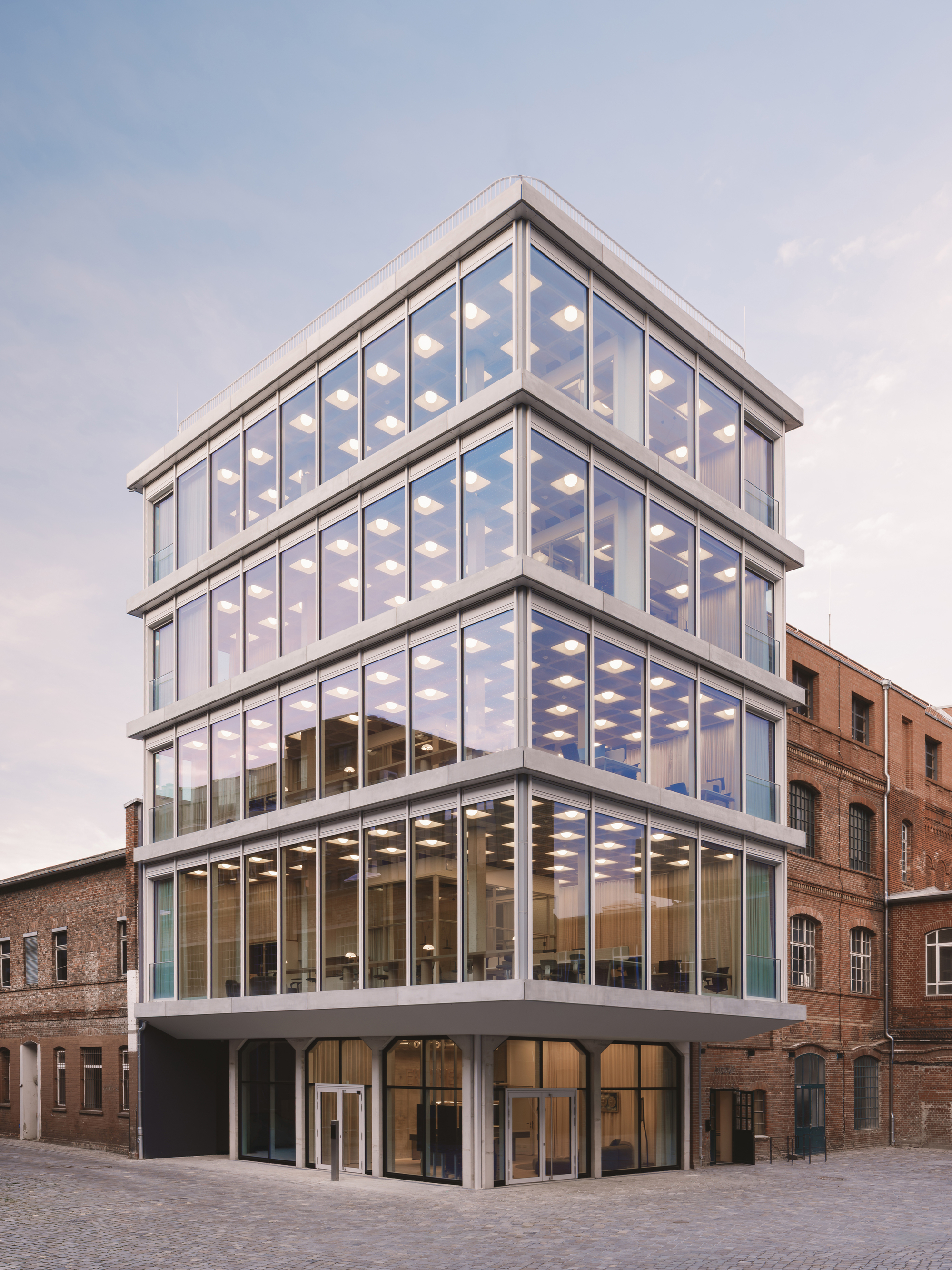 Corner view of the stacked glass facade with illuminated ceiling grids at dusk beside brick buildings