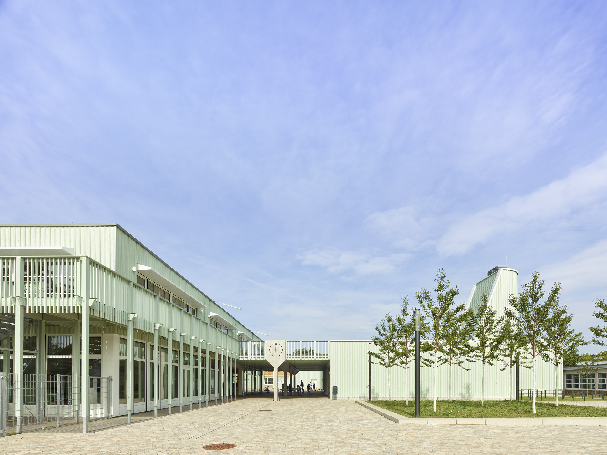 Paved courtyard flanked by pale green corrugated metal facades with white columns and young trees