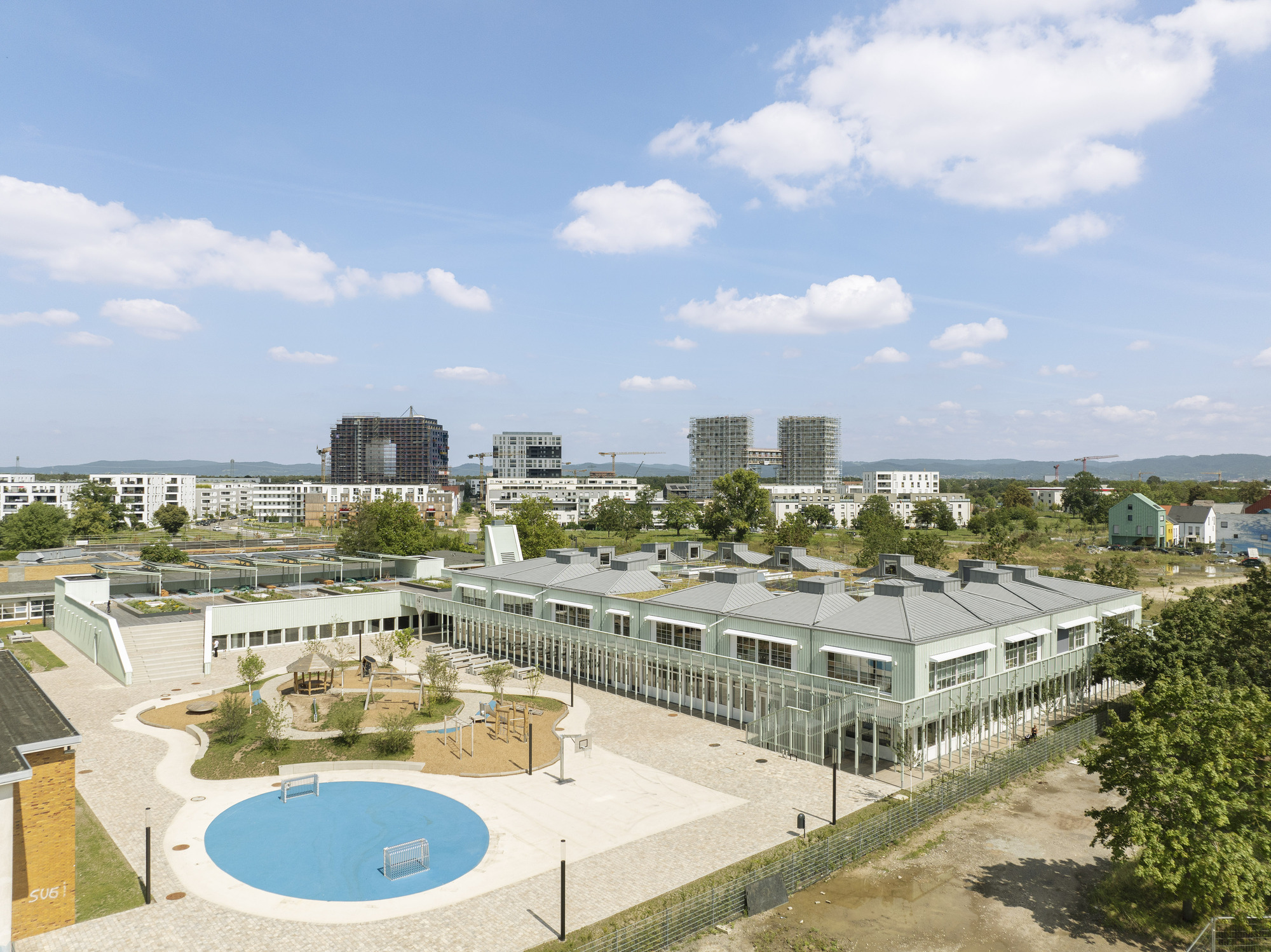 Aerial view of the pale green corrugated roofscape with circular blue pool in the foreground plaza