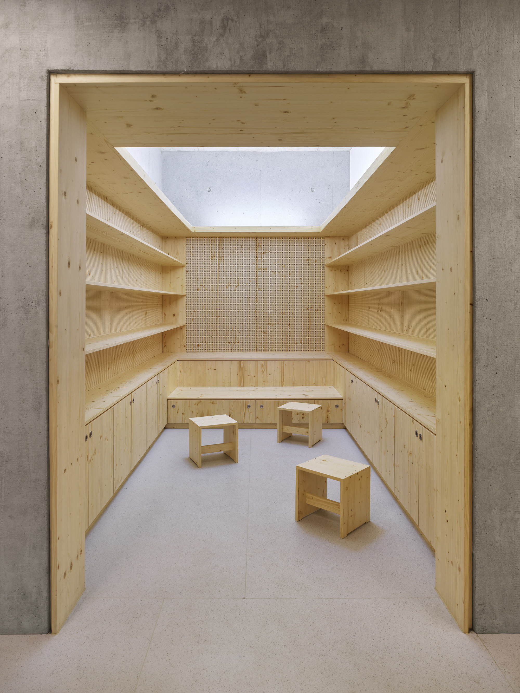 Pine shelving alcove framed by concrete walls with a central skylight above three stools
