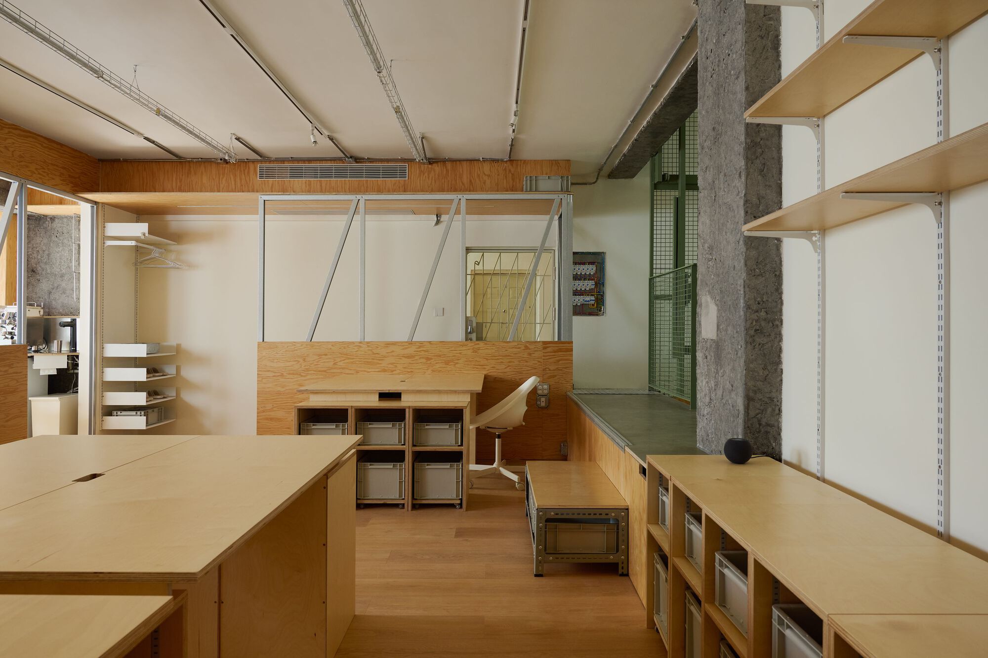 Interior workspace with plywood workbenches and exposed concrete columns beneath track lighting and a dropped ceiling