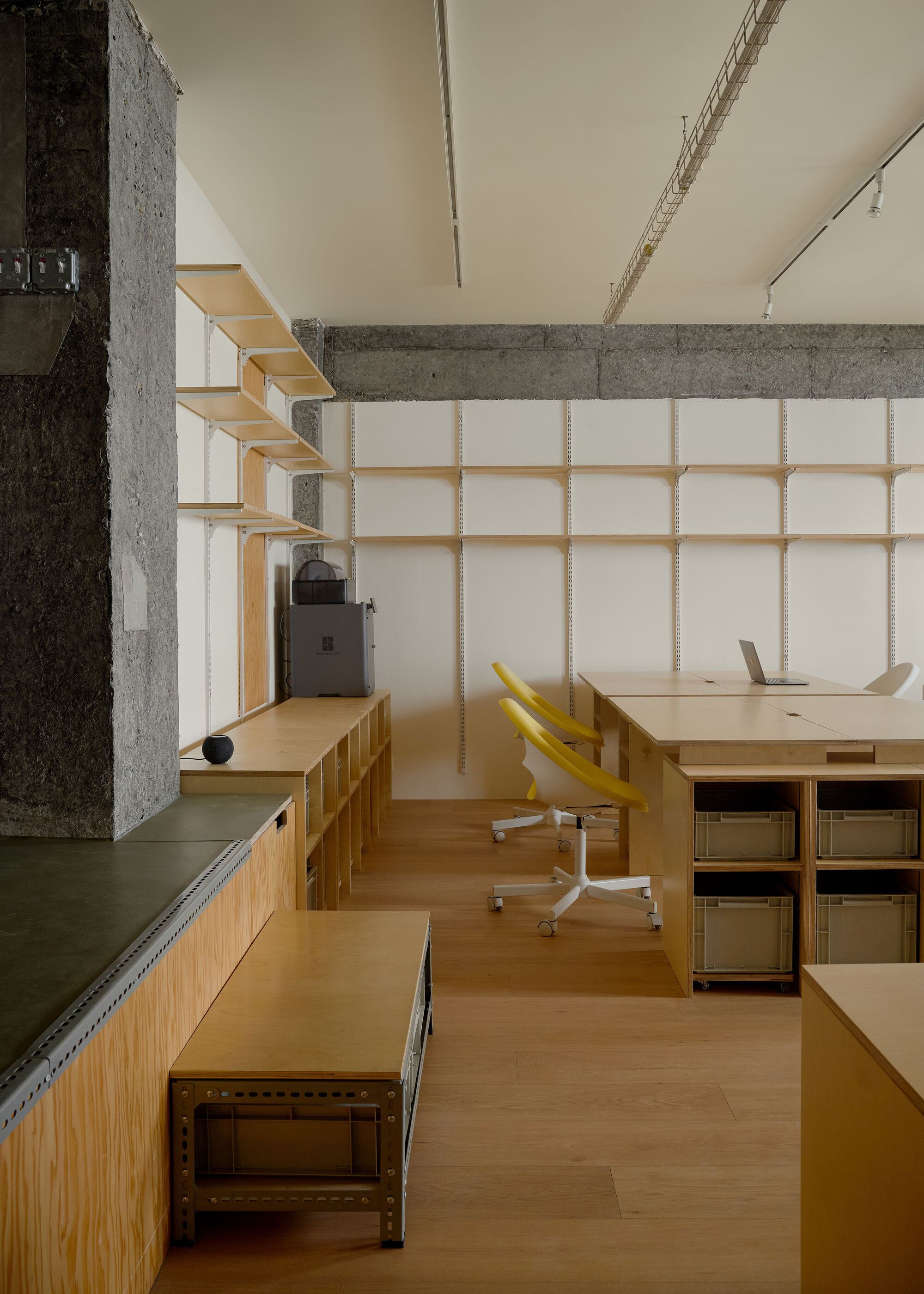 Corner workspace showing plywood desk with yellow chair facing gridded translucent wall panels under concrete beams