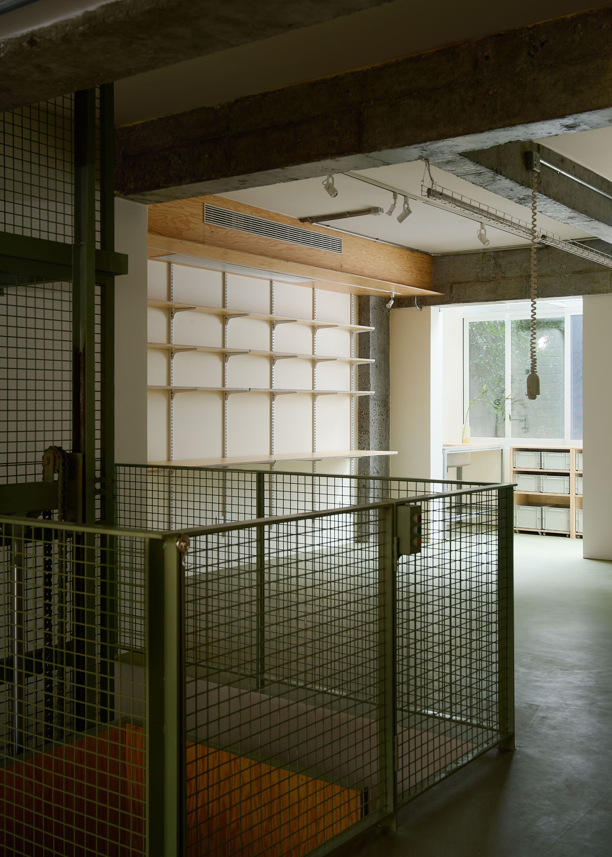 Interior view through green wire mesh partitions toward timber-framed shelving beneath exposed concrete beams