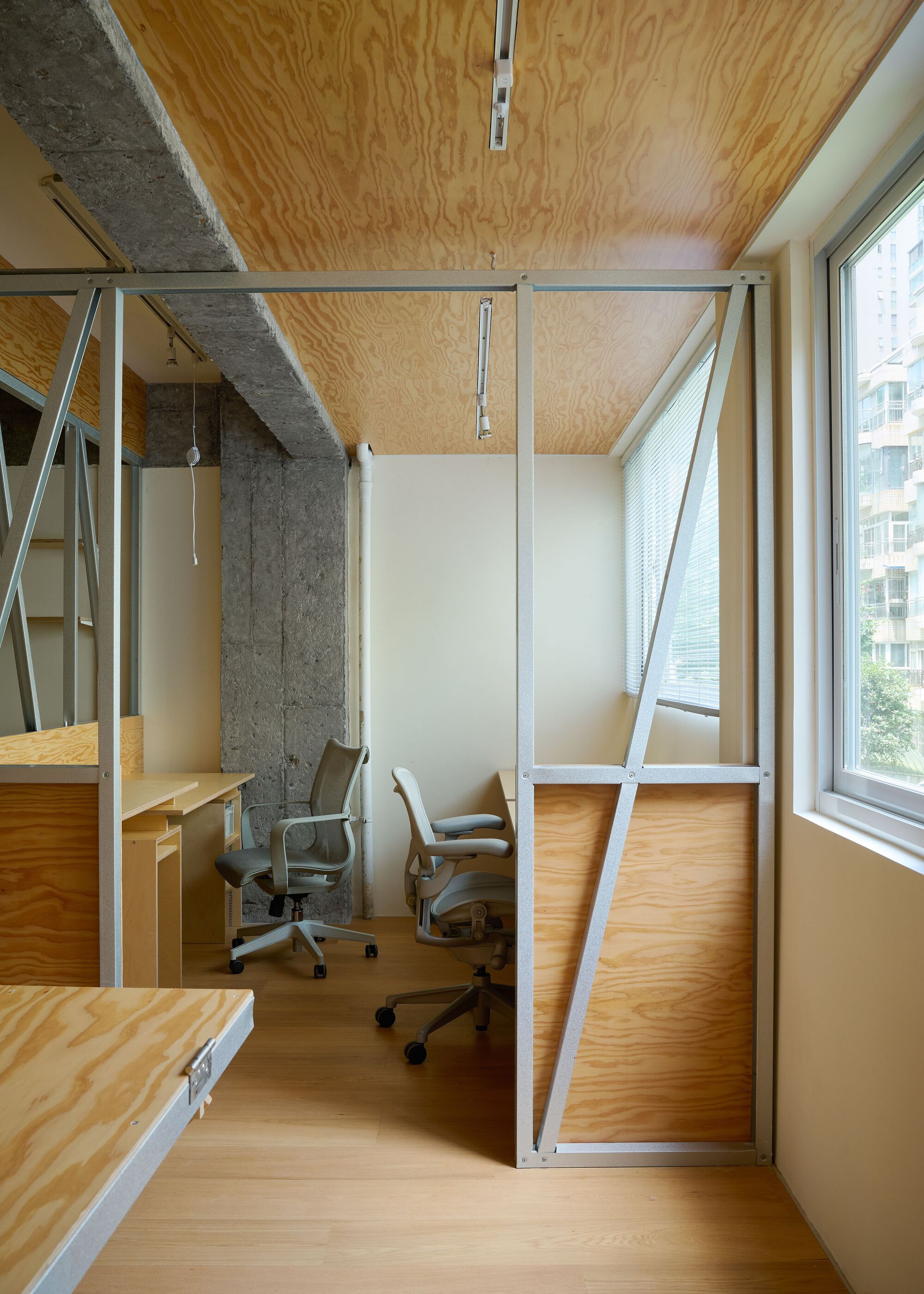 Enclosed workspace cubicle with steel framing and plywood paneling beneath a plywood ceiling and track lighting