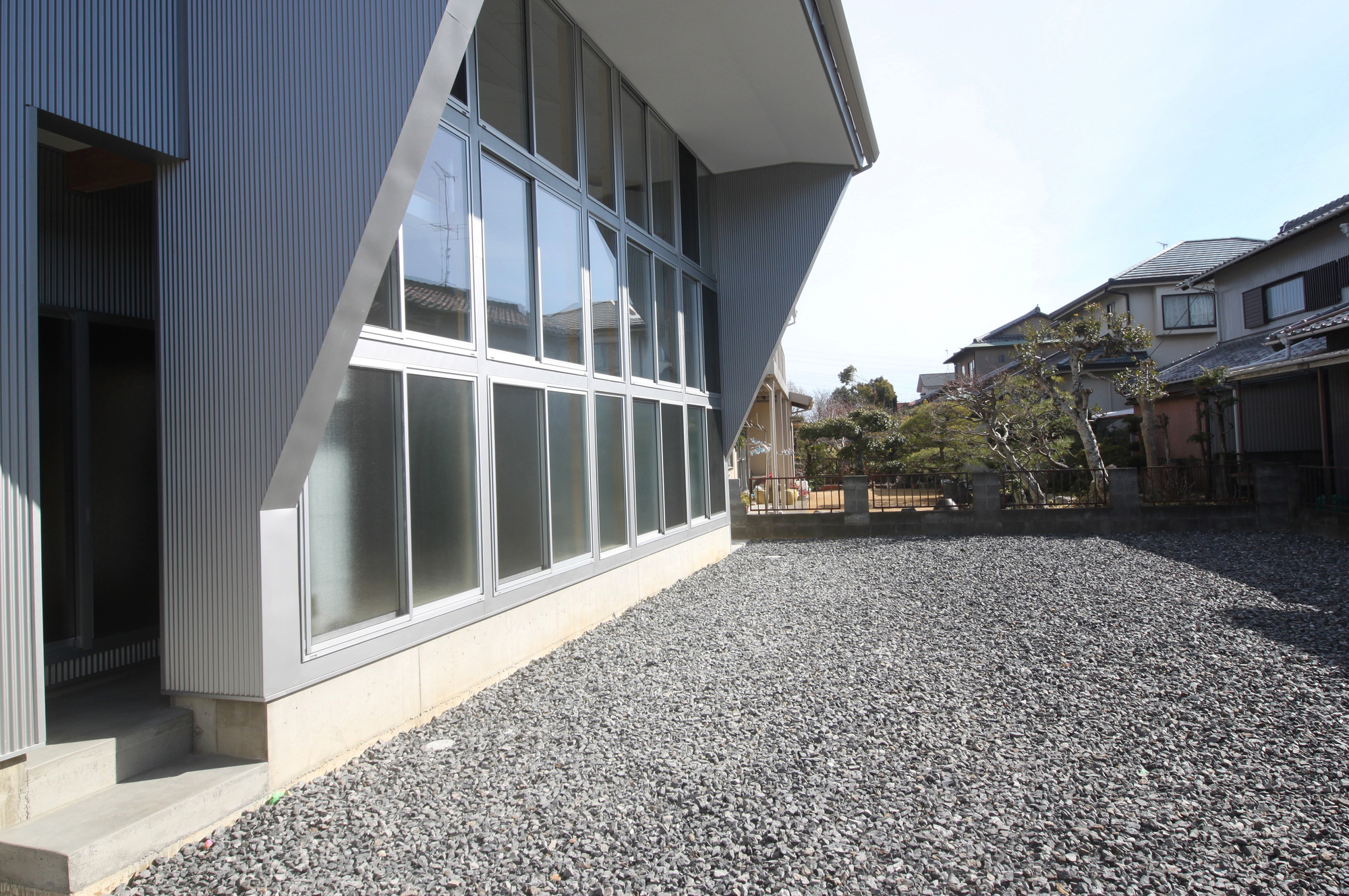 Corrugated metal facade with angled soffit above full-height glazing facing gravel yard in daylight