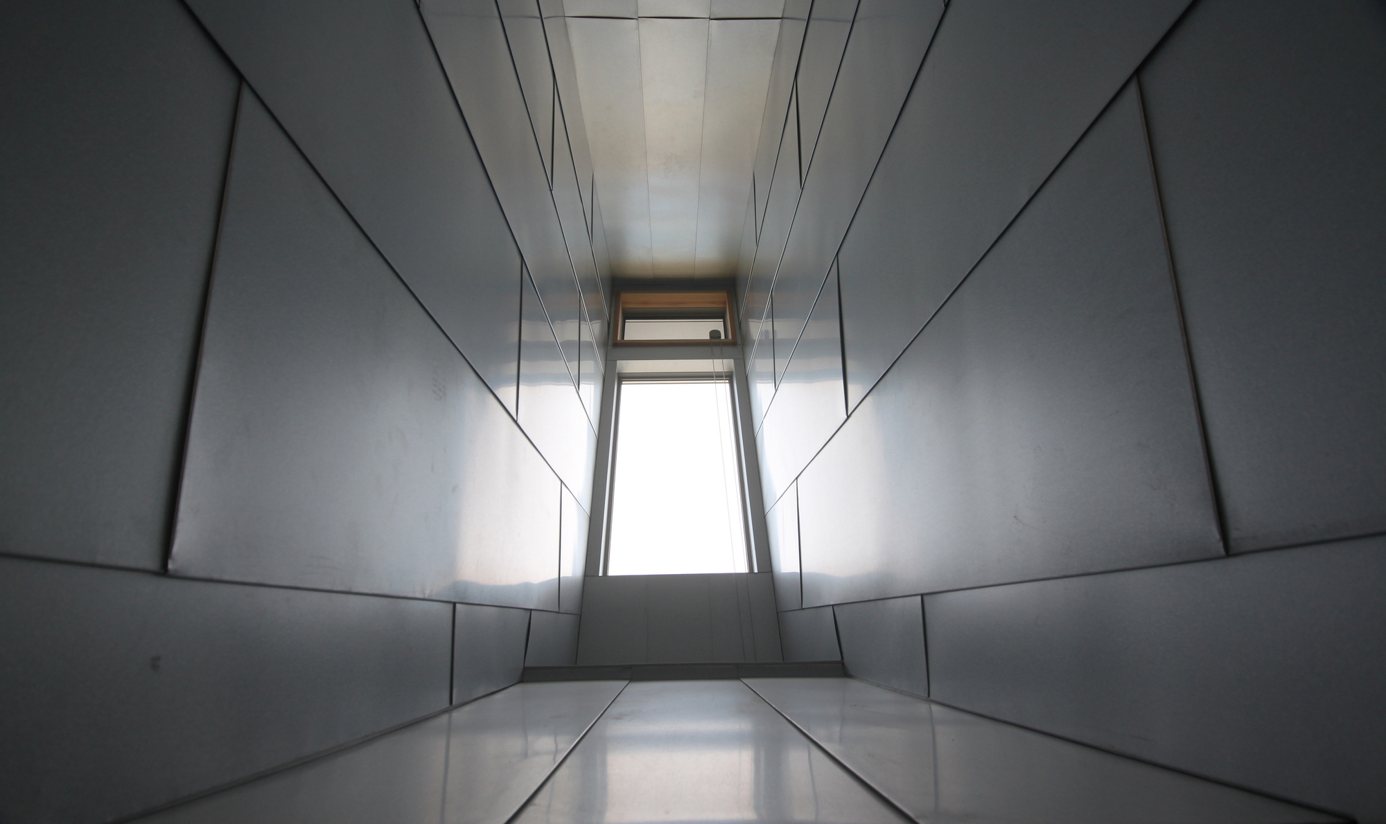 Narrow corridor lined with metal panels converging toward a distant window at the end