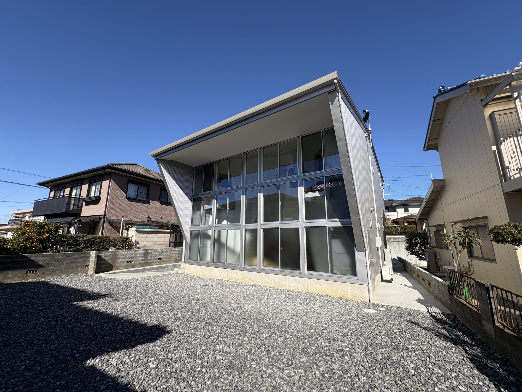 Two-story glass and metal facade with canted roof plane set in a gravel yard under clear blue sky