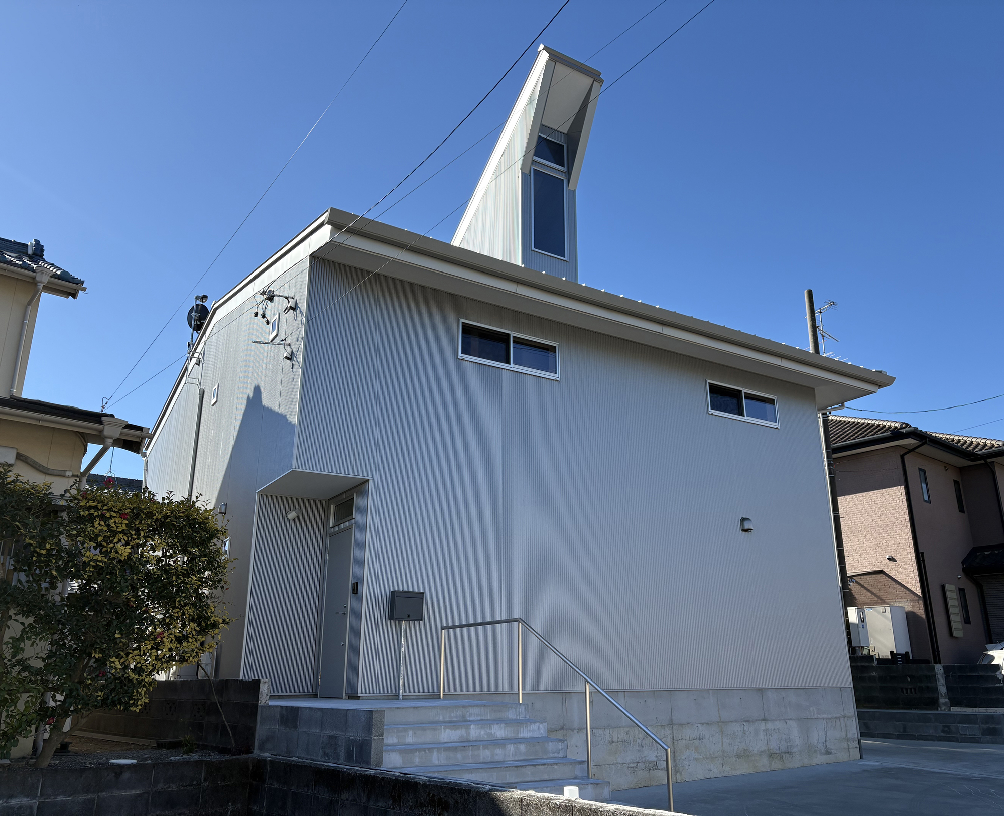 Corrugated metal exterior with vertical tower element and concrete entrance stairs among neighboring houses