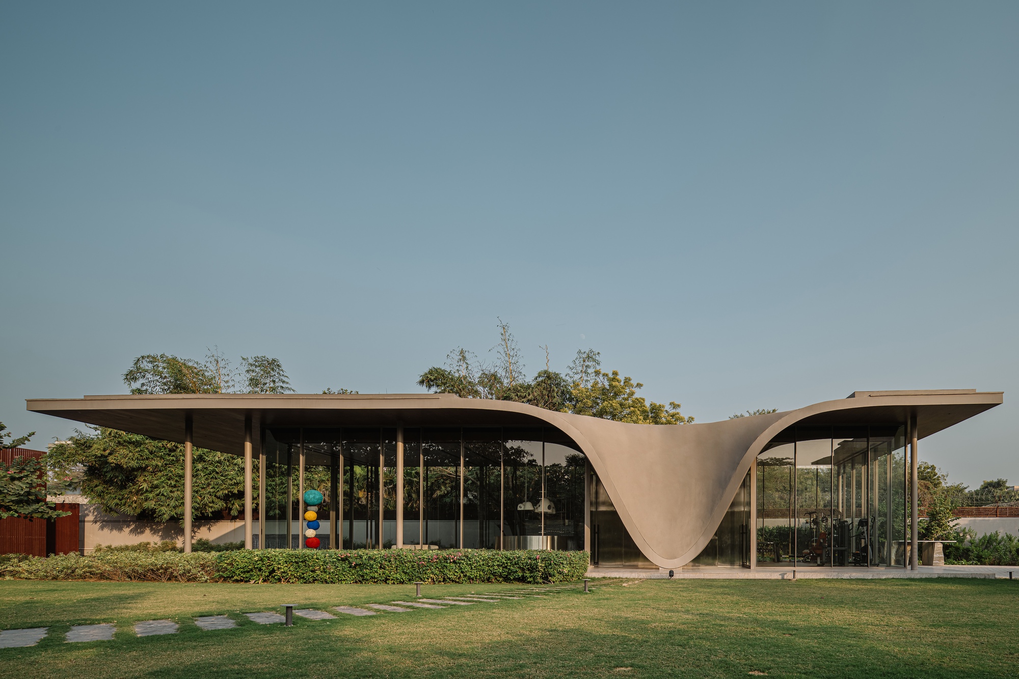 Glass pavilion with flowing concrete roof canopy set in a lawn with hedges and trees