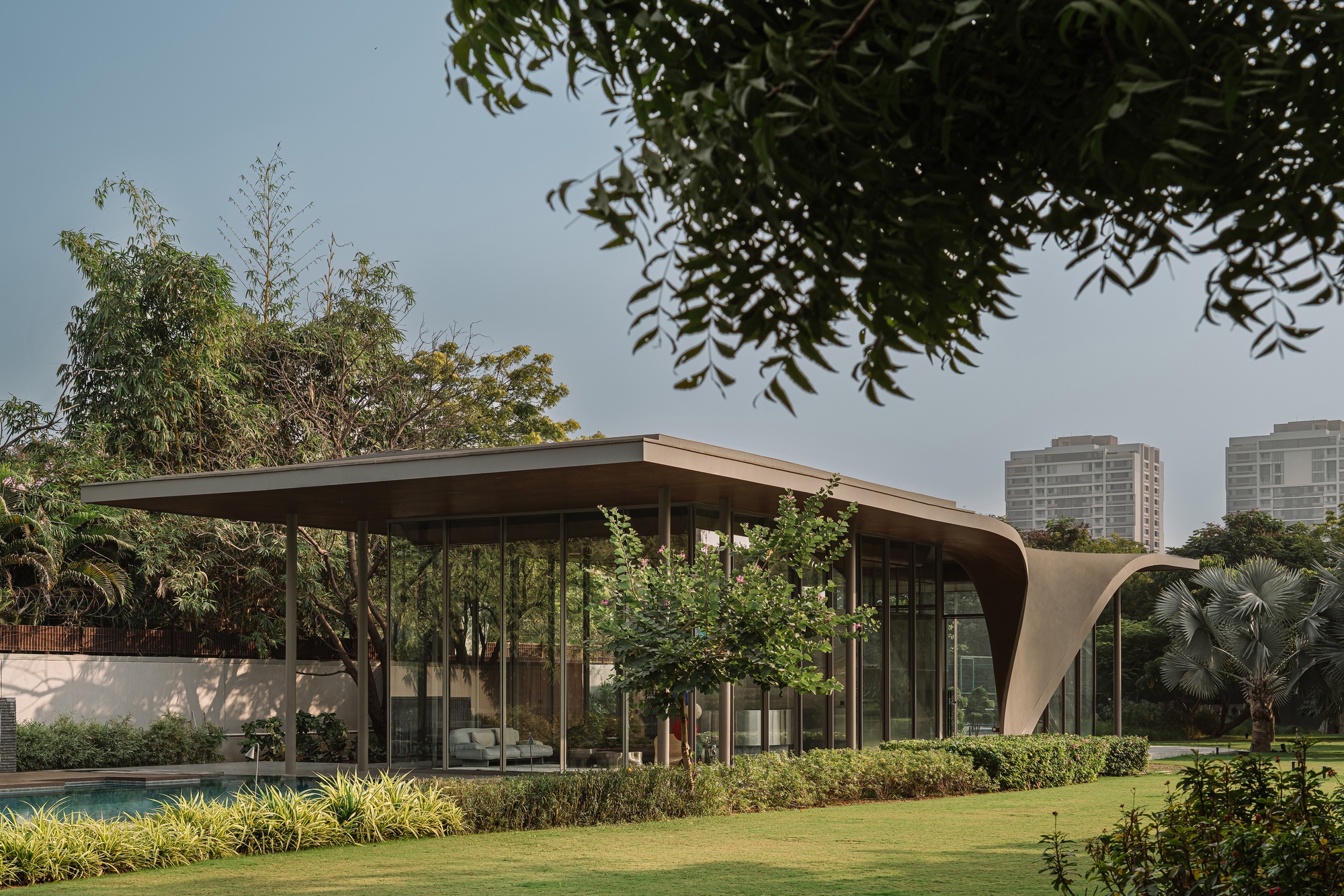 Side view of the glazed facade beneath overhanging roof planes framed by garden foliage