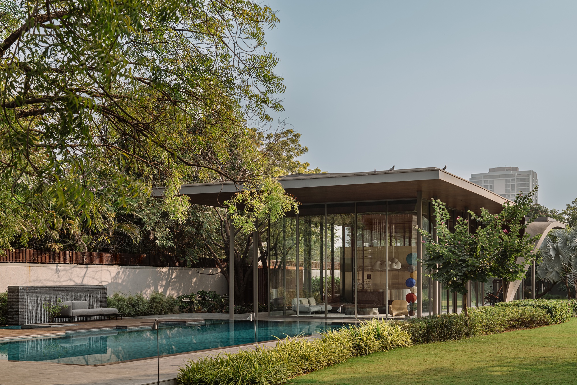 Glass pavilion with flat roof and slender columns beside a reflecting pool and lawn with mature trees