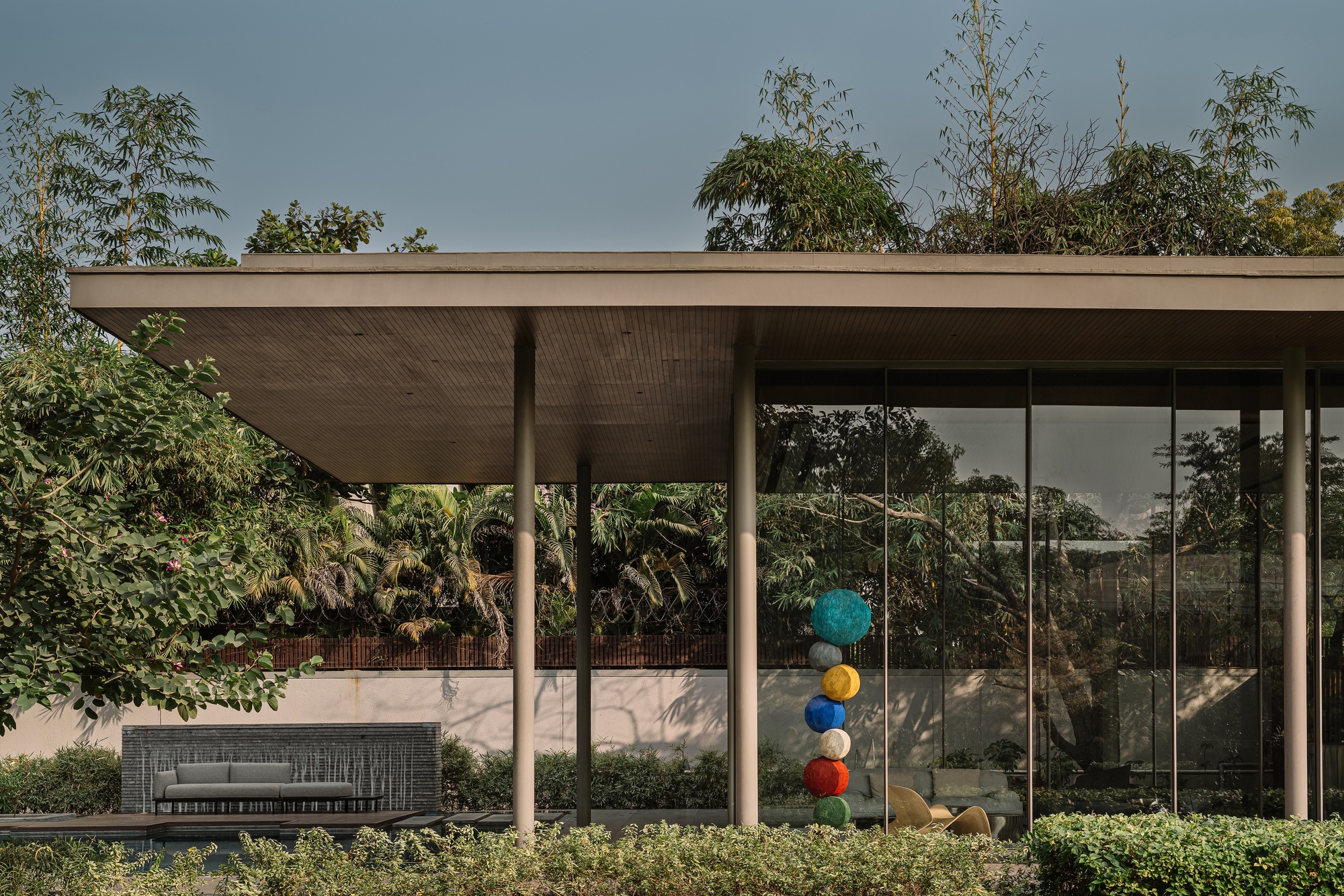 Covered terrace with timber-lined soffit and glass walls framing a sculpture beneath an overcast sky